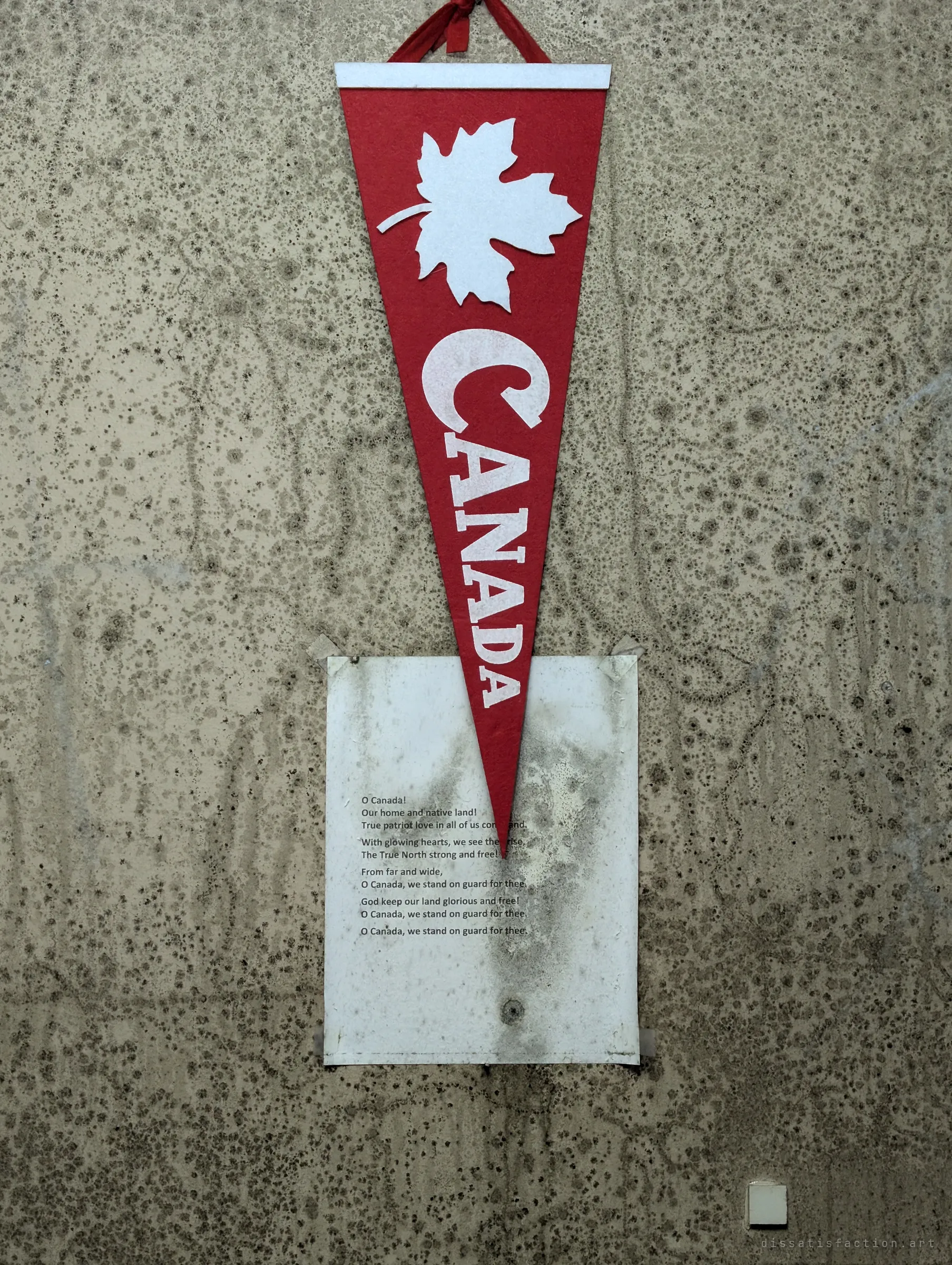 A red felt pennant with a maple leaf hangs over a printed copy of the national anthem, both taped to a wall speckled with mould. The patriotic decoration is slowly being eaten by the damp.