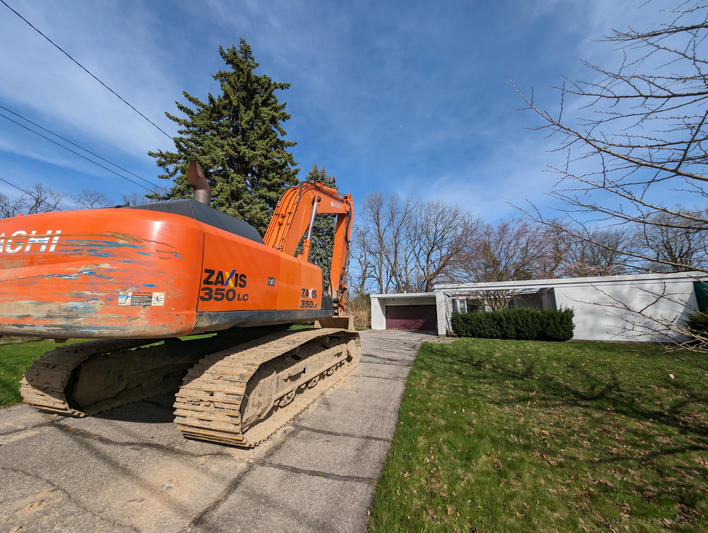 A mud-caked orange excavator sits on the driveway in front of a low white flat-roofed house, a sign the place is about to be torn down.