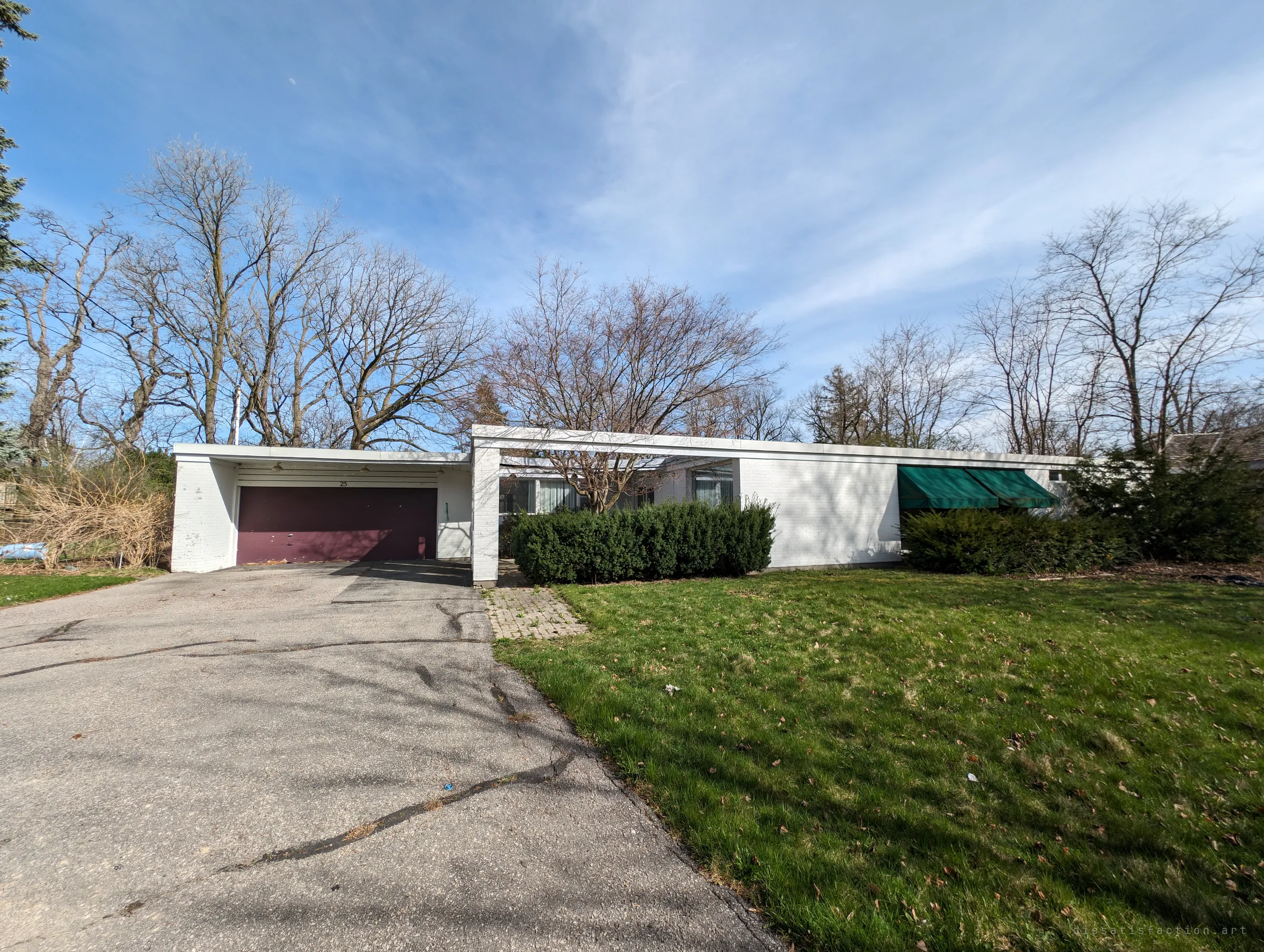 The long, low white brick house seen head-on from the driveway, its flat roof and carport framed by bare spring trees and an overgrown lawn.