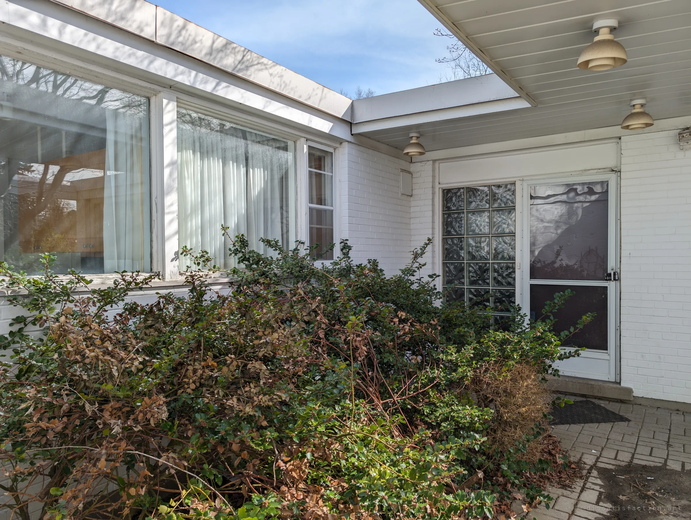 An overgrown shrub swallows the covered entry, with glass-block panels and a locked storm door under mushroom-shaped porch lights.