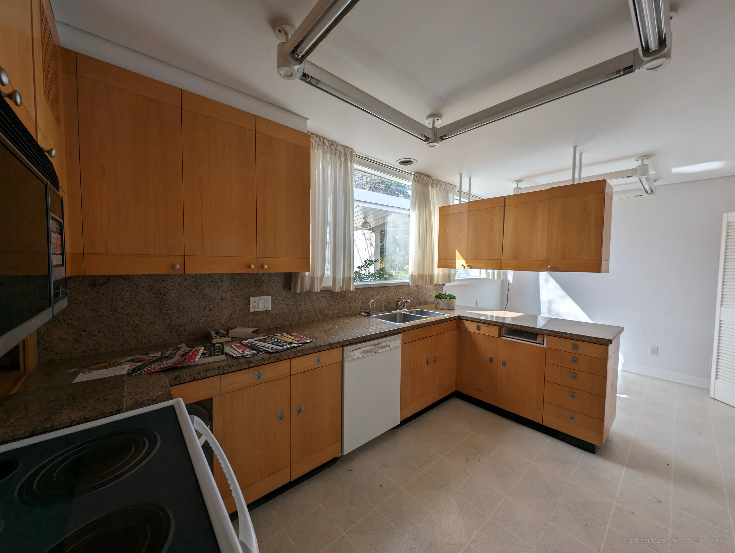 The whole kitchen from the stove: matching wood cabinets wrap around the room under suspended fluorescent light rails, sunlight falling across the empty floor.