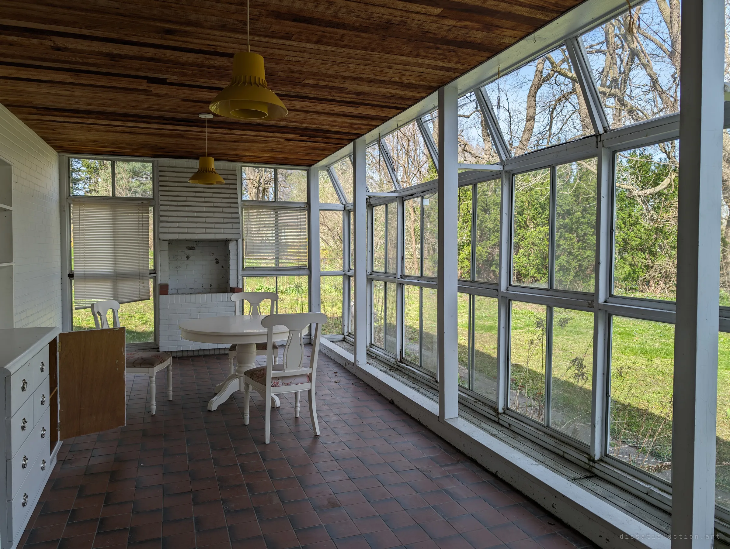 A glass-walled sunroom with a wood-plank ceiling, yellow pendant lamps and a white table set, looking out onto an overgrown backyard.