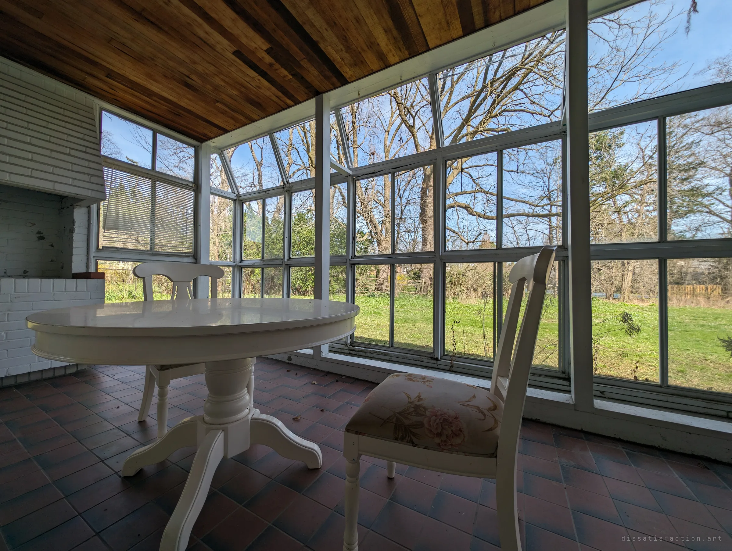 Low angle across the sunroom table, the slanted glass walls filled with bare trees and a bright green lawn gone wild.