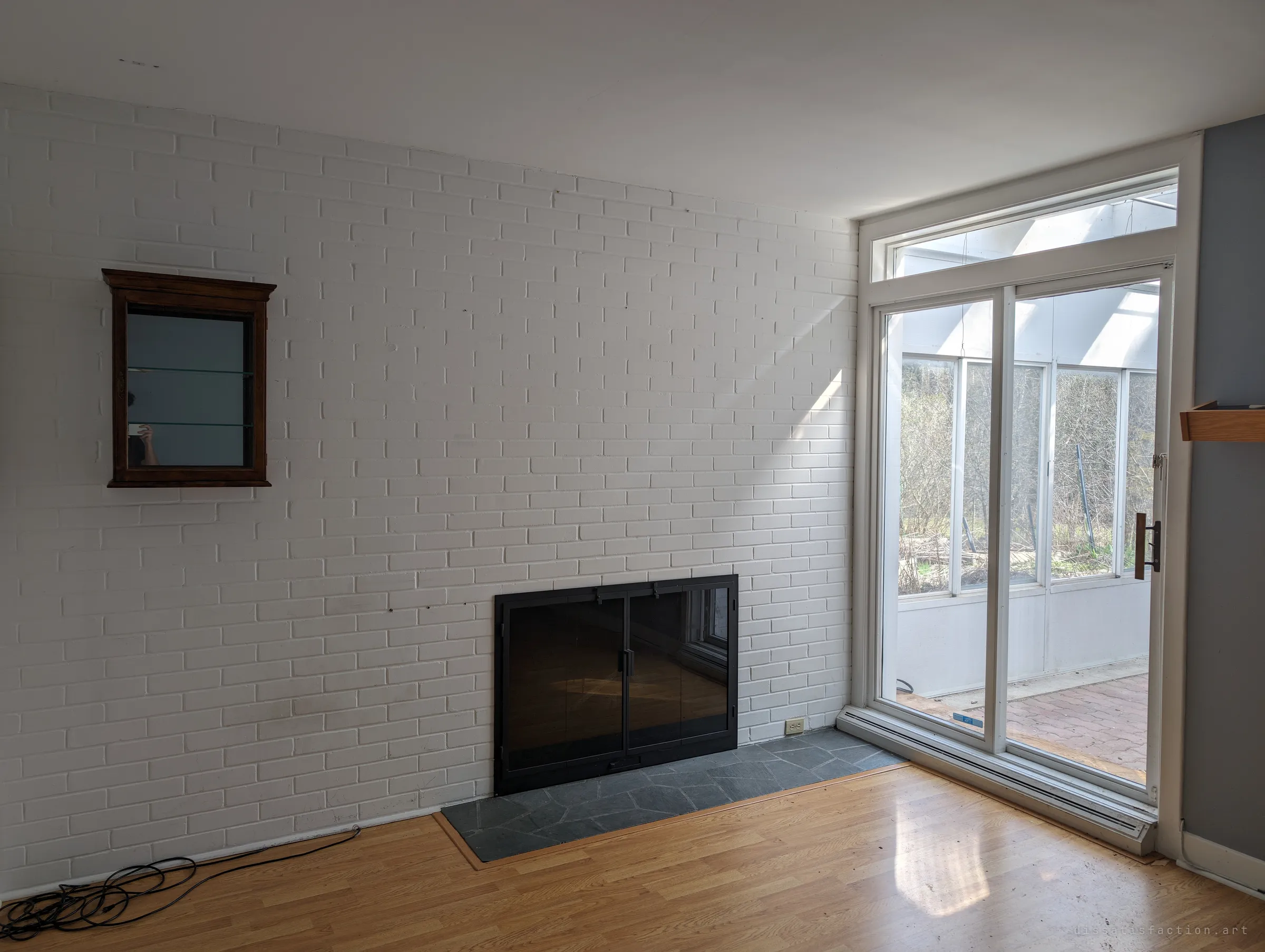An empty living room with a painted white brick wall, a black-doored fireplace and a small mirrored cabinet, sliding doors opening onto the sunroom.
