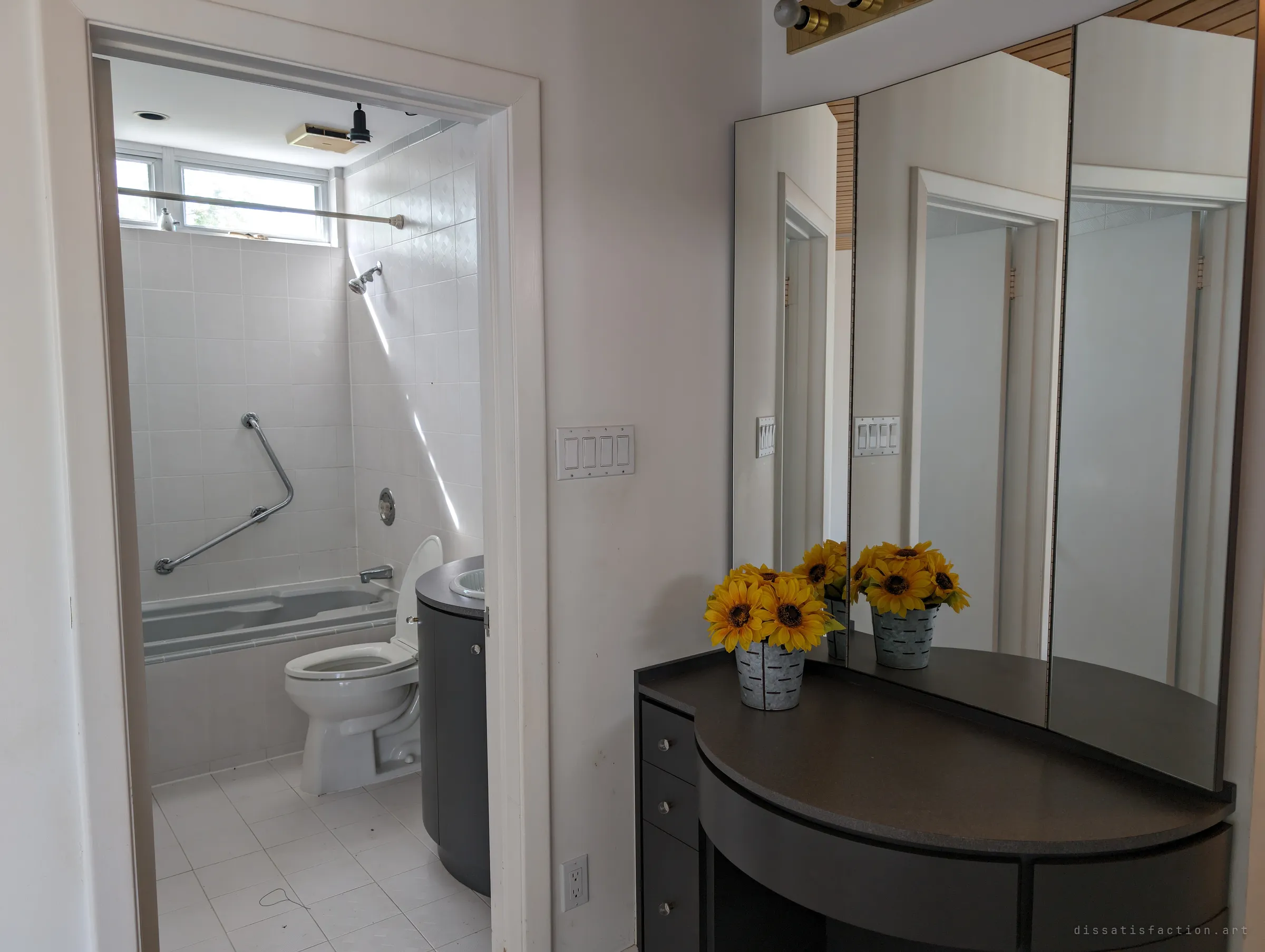 A dressing area with a curved dark vanity, a tri-fold mirror and a pot of artificial sunflowers, opening onto a white-tiled bathroom lit by a clerestory window. The staged flowers are an odd touch of life in an otherwise emptied house.
