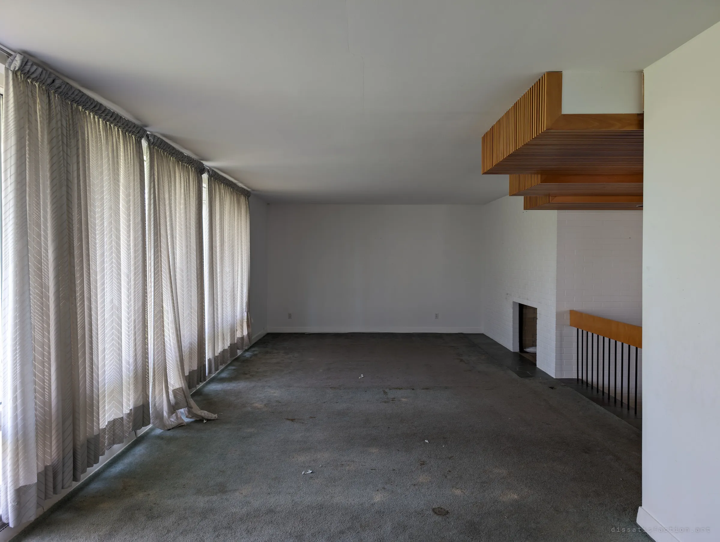 An empty living room with a wall of sheer floor-length curtains, worn green carpet, a white-painted brick fireplace and a slatted wood ceiling feature over a sunken stair. The mid-century details survive while the room itself has been stripped bare.
