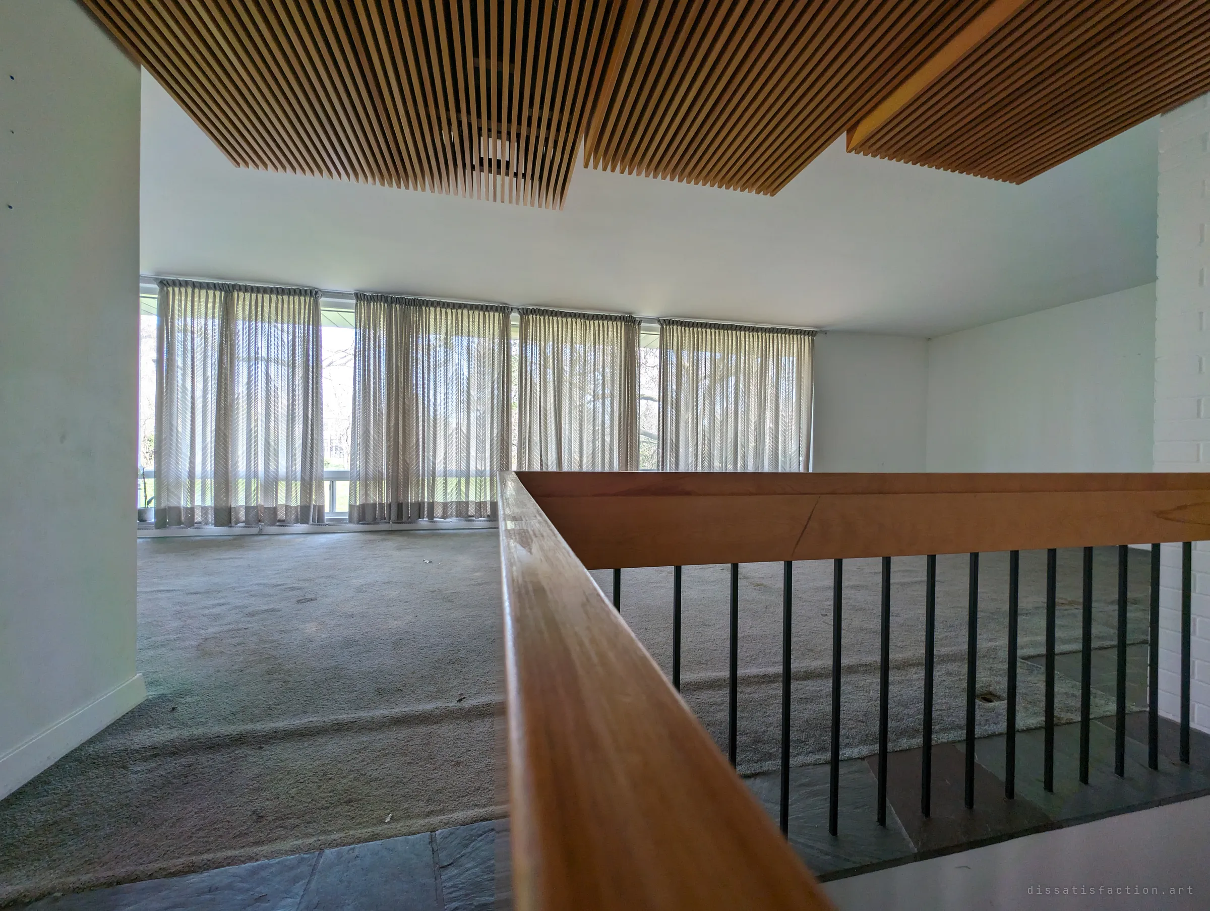 Looking past a wooden handrail and dropped slat ceiling toward a long wall of curtained windows, with bare trees glowing behind the fabric. The low angle shows the split-level layout of the main living space.