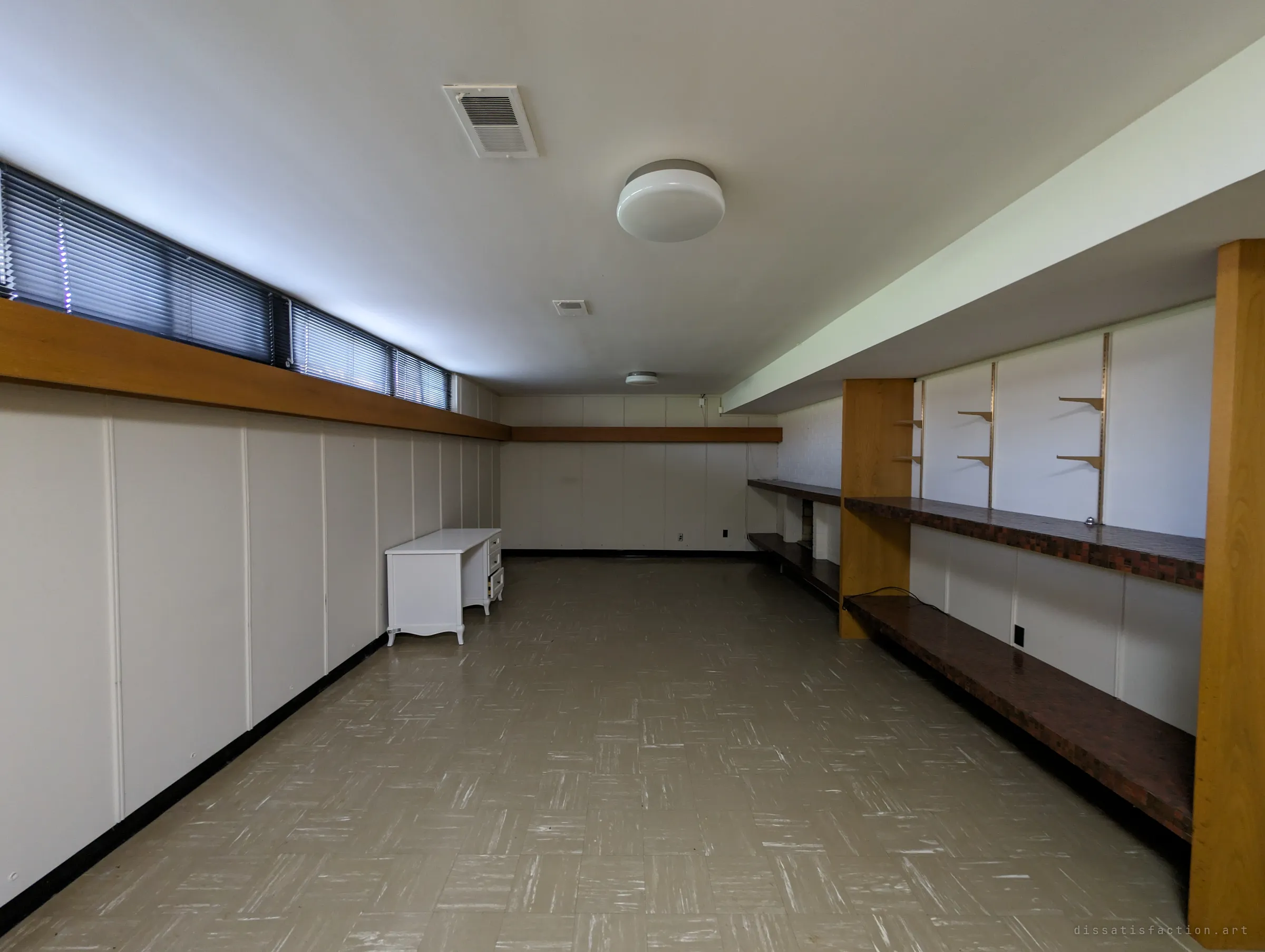 A long basement rec room with panelled walls, a band of high windows behind blinds, built-in tiled counters and empty adjustable shelving, and a lone white desk. The empty shelves hint at a collection that once filled the room.