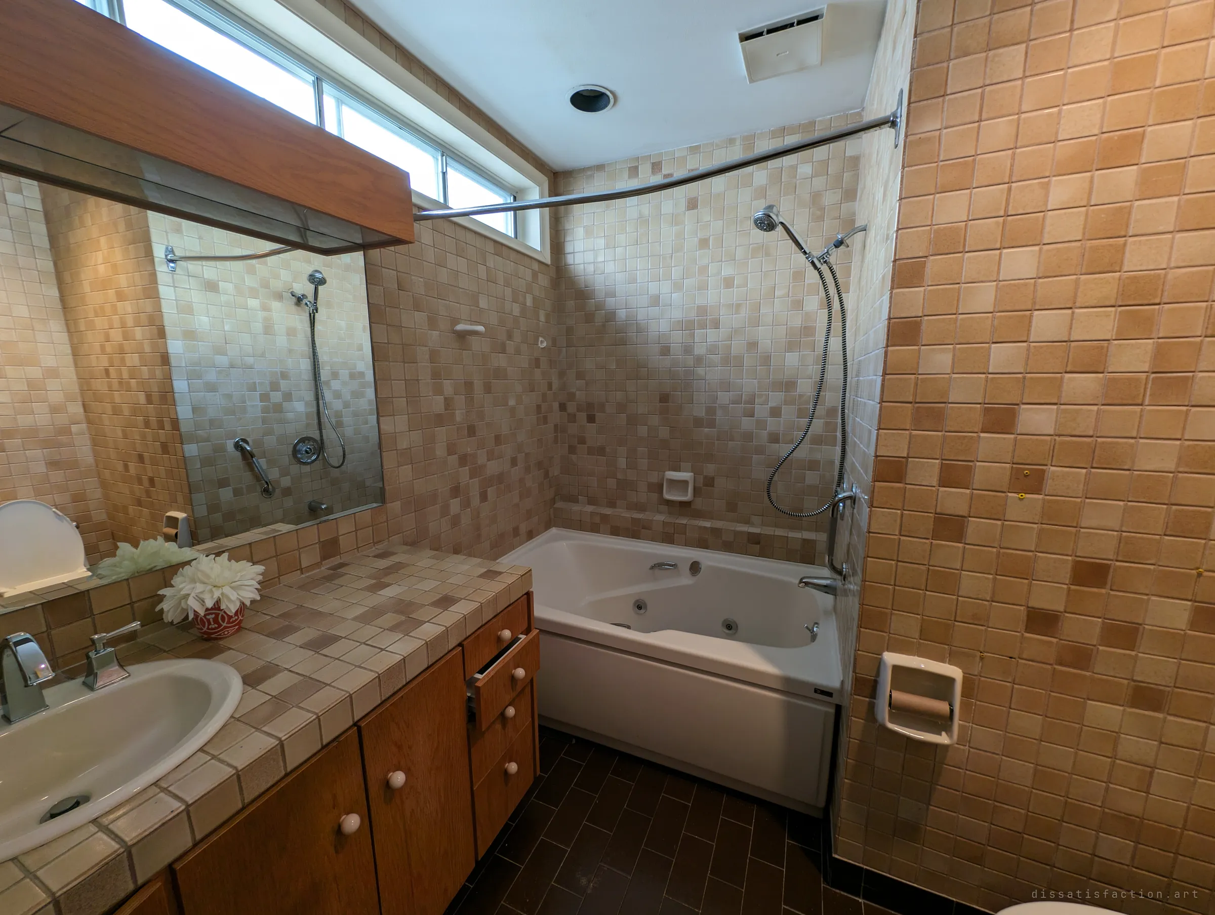 A bathroom wrapped floor to ceiling in warm beige mosaic tile, with a jetted tub, a handheld shower and a tiled vanity reflected in a wide mirror. The original tilework is a well-preserved slice of the house's era.