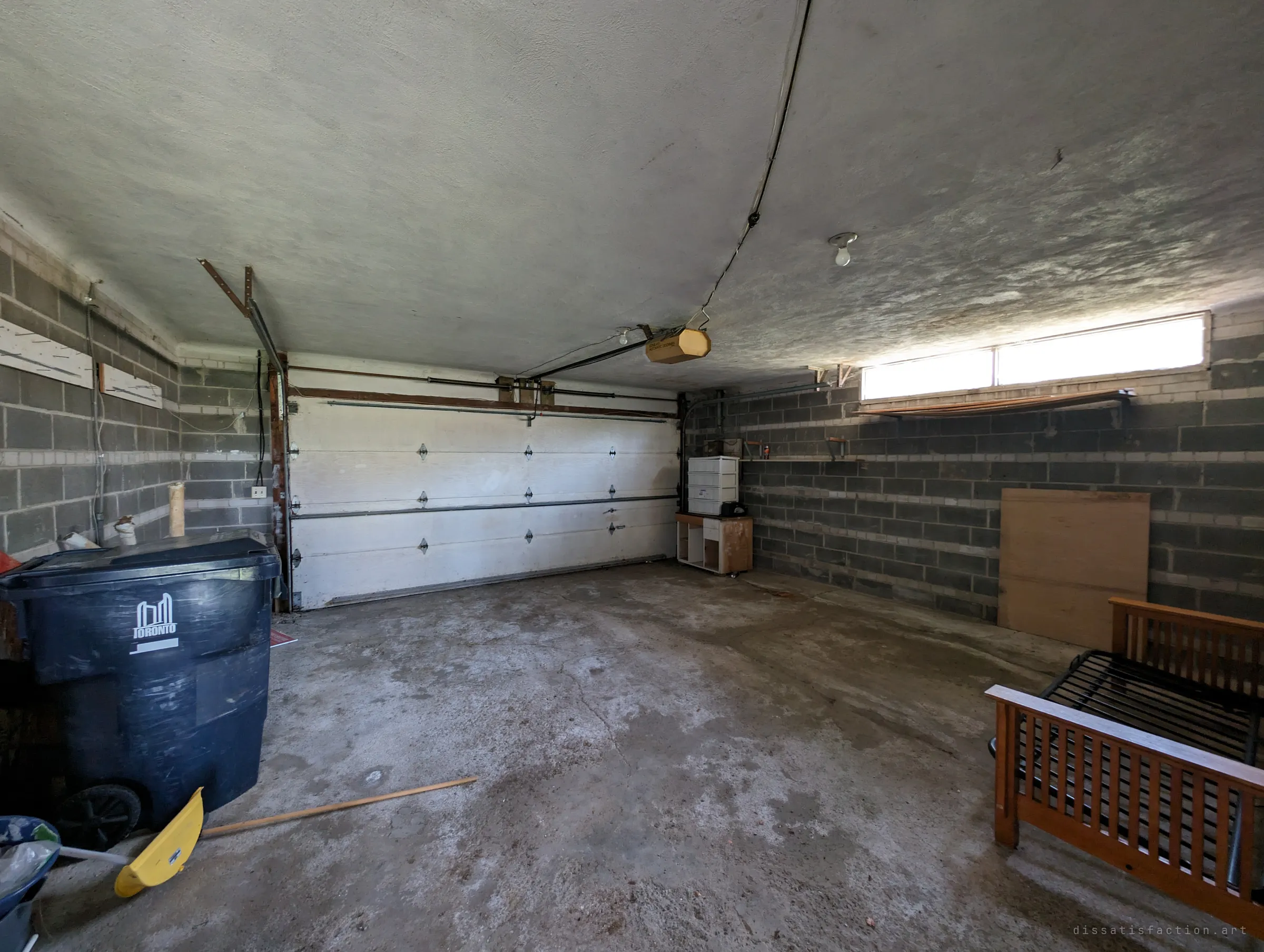 A cinder-block garage with a closed white overhead door, a large wheeled bin, a snow shovel on the concrete floor and a wooden futon frame leaning by the wall. Daylight from a single strip window is all that lights the space.