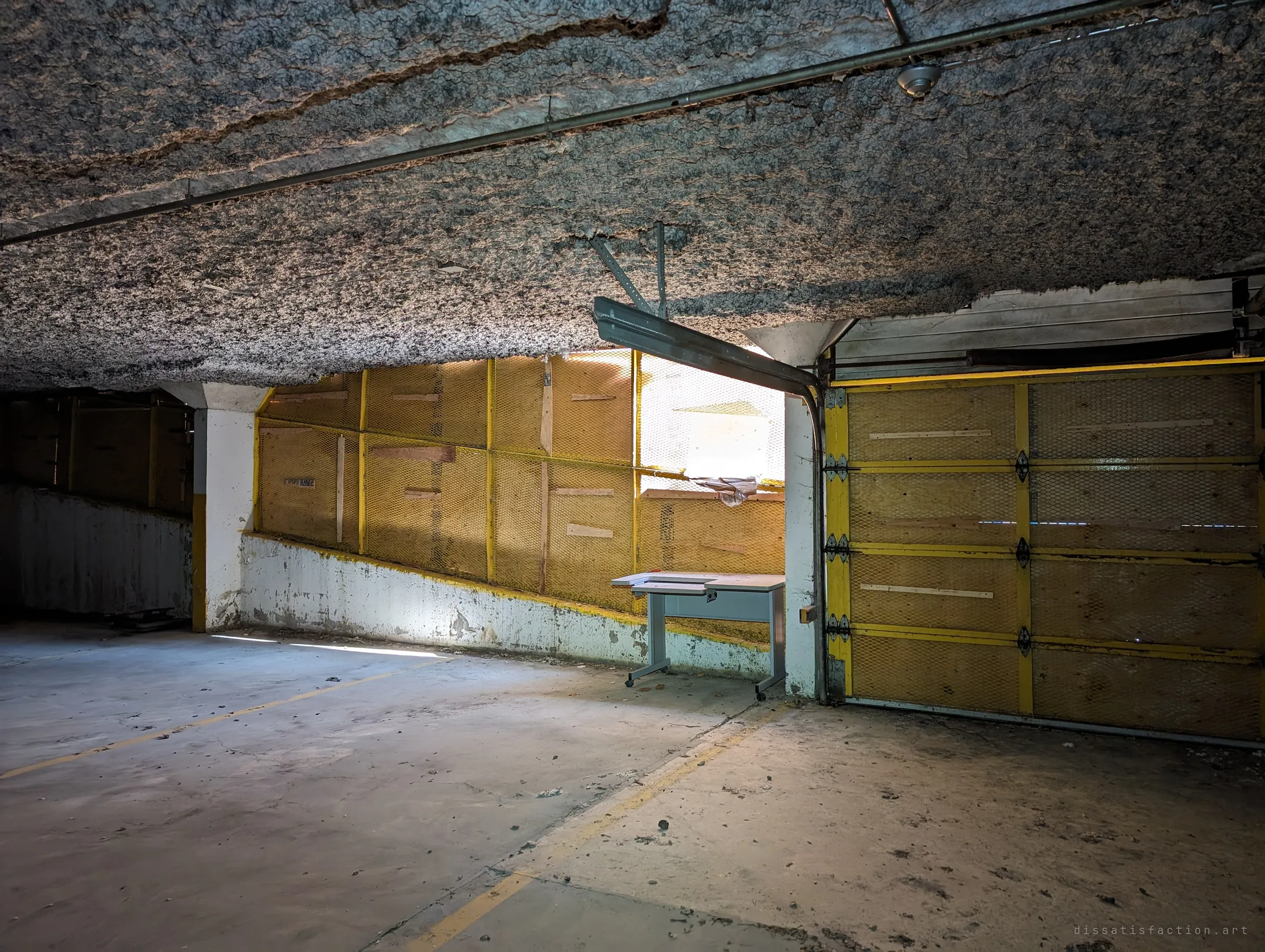 Inside the parking garage, a sagging insulated ceiling hangs over yellow-trimmed boarded-up openings and a lone desk left by the ramp. Daylight leaks in only through the mesh-covered gaps.