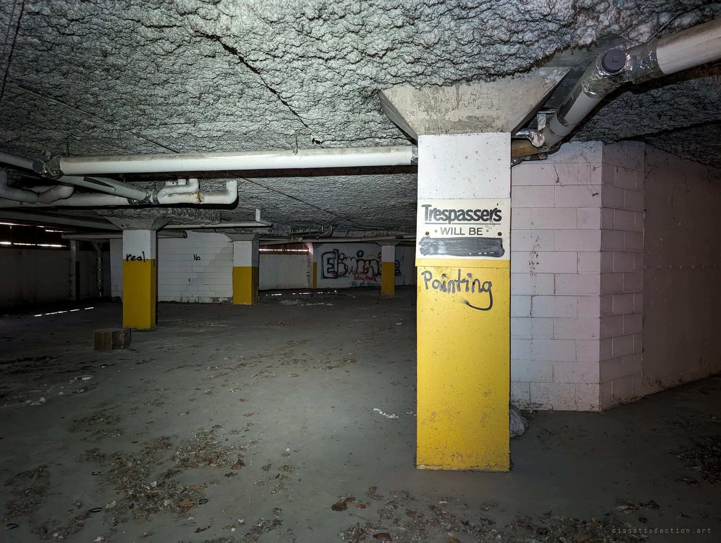 A yellow and white pillar carries a trespassing sign with its last word painted over and scrawled into a joke, deep in the dark underground garage. Graffiti and more pillars fade into the black behind it.