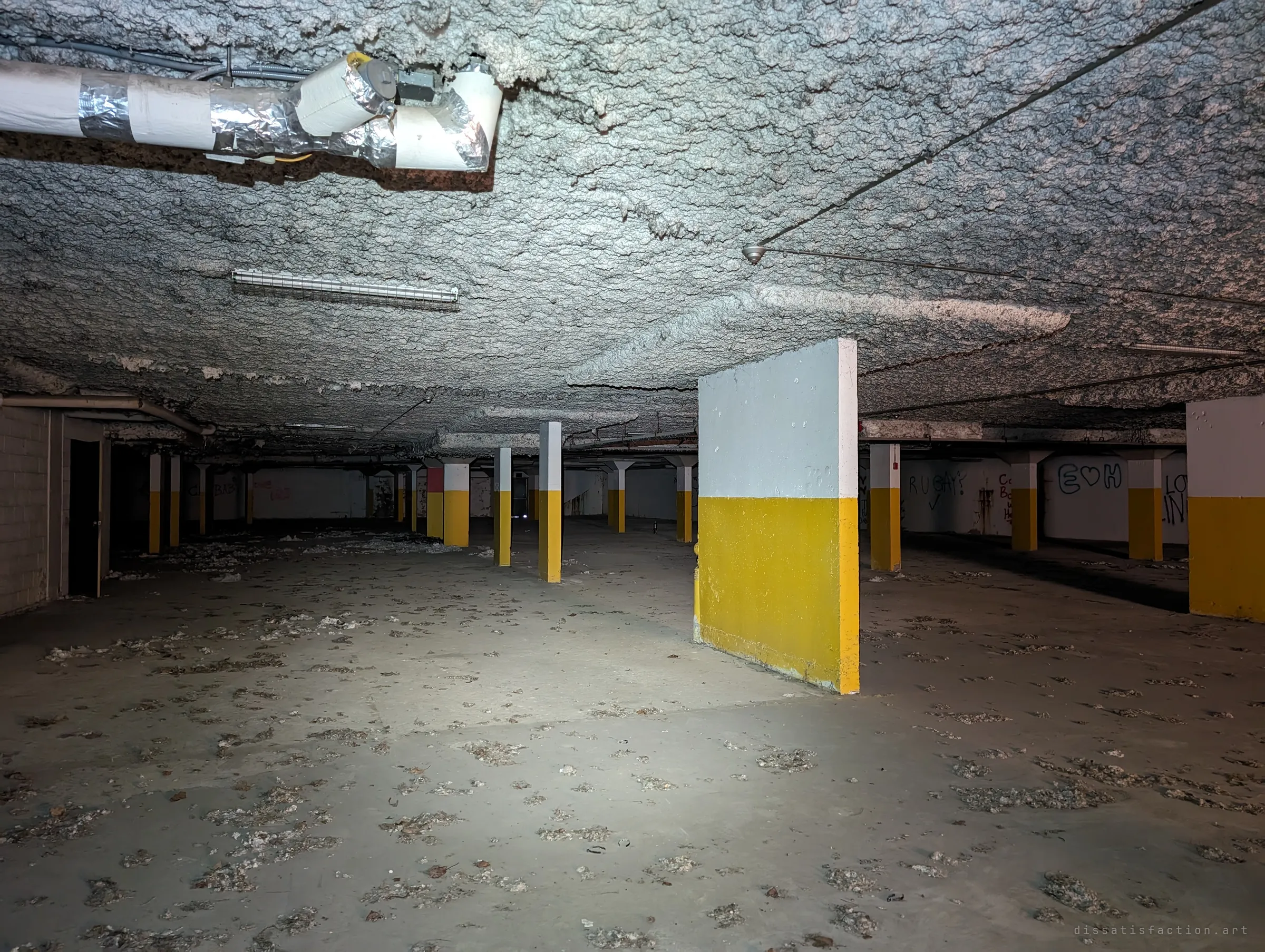 A flashlight picks out a painted concrete wall stub in the empty garage level, with rows of yellow-banded pillars running into the dark. Clumps of fallen ceiling insulation litter the floor.