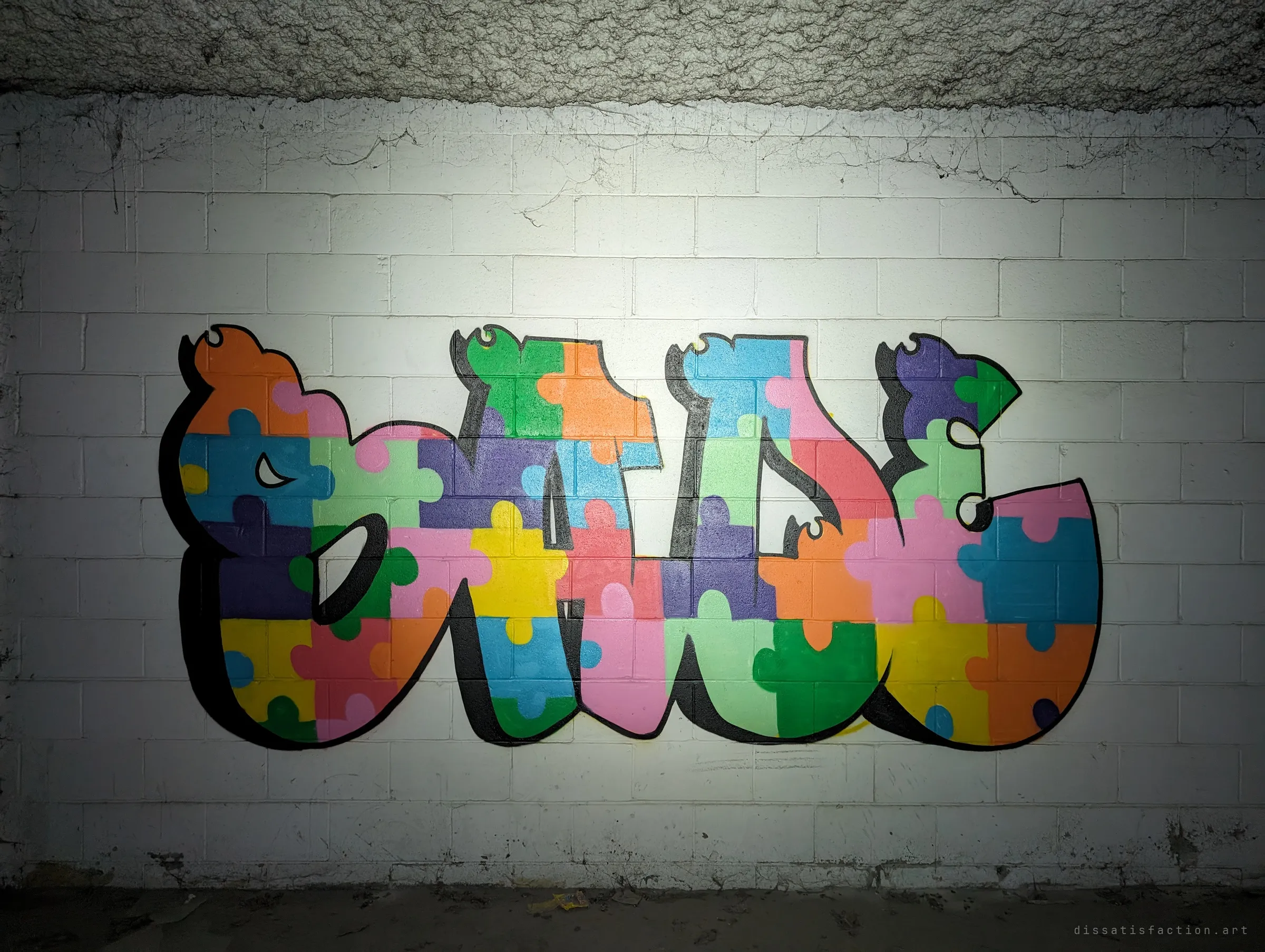 A bright graffiti piece filled with multicoloured jigsaw pieces covers a whitewashed block wall under the garage ceiling. It is one of the cleanest pieces left in the dark.