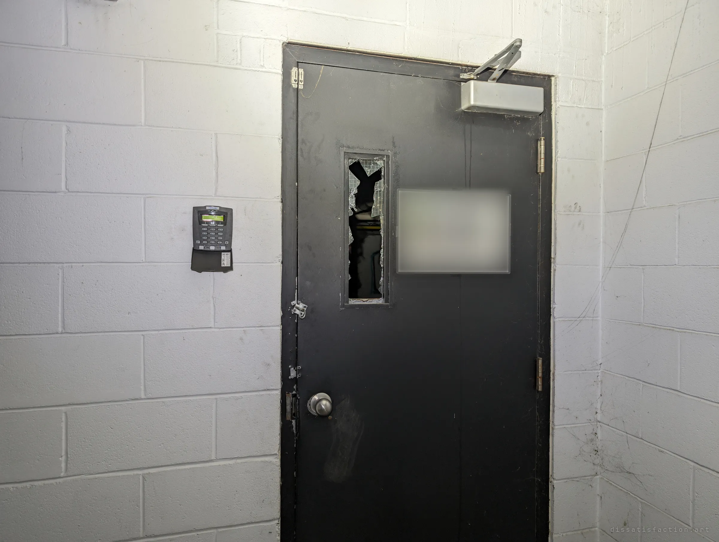 A black steel door with a smashed wired-glass window and an old access keypad on the block wall beside it. This was the way in from the garage to the stairs.