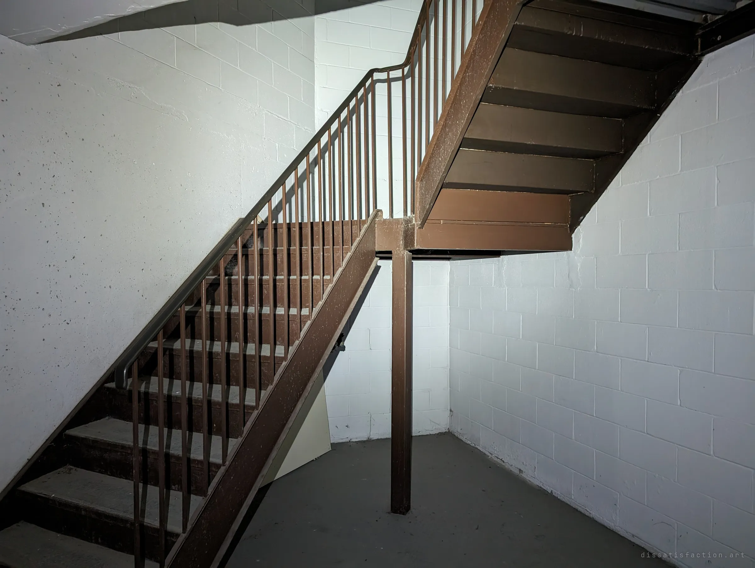 A brown steel staircase with thin balusters climbs through a whitewashed block stairwell. The stairs lead up from the garage into the building.