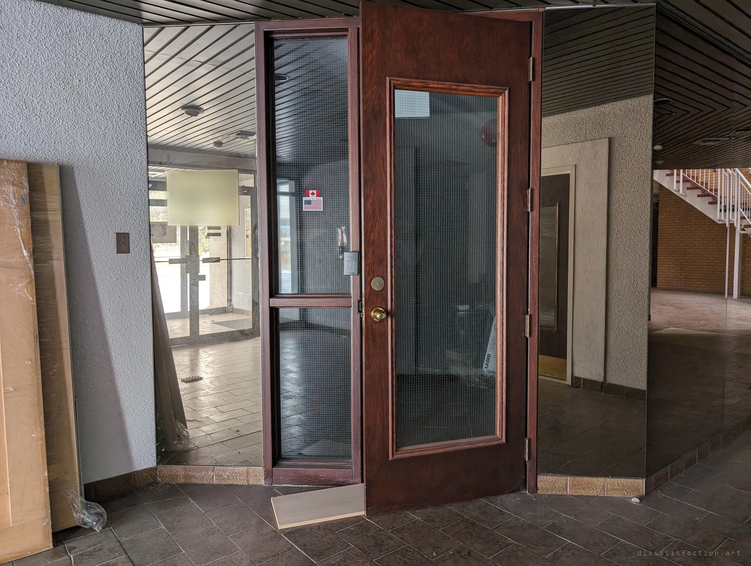 A wood-framed glass door stands propped open onto a mirrored lobby with dark slatted ceilings and a reception sign behind glass. The lobby still looks ready for visitors that never come.