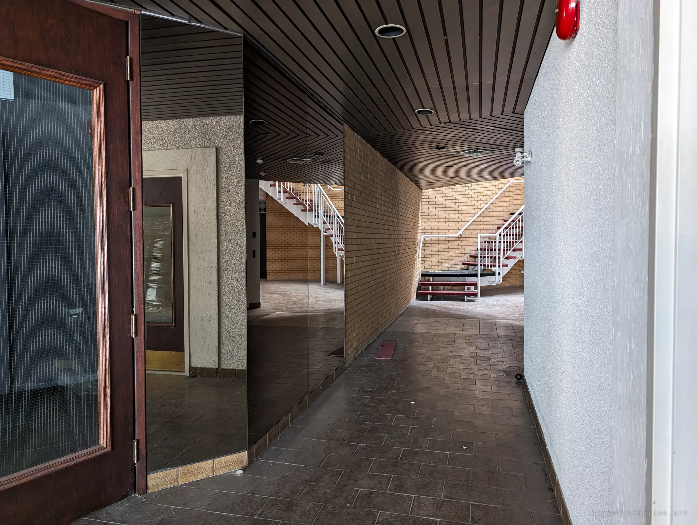 A tiled corridor under a dark slatted ceiling runs past mirrored walls toward a yellow-brick atrium with red-treaded white stairs. The mirrors double the empty hallway.