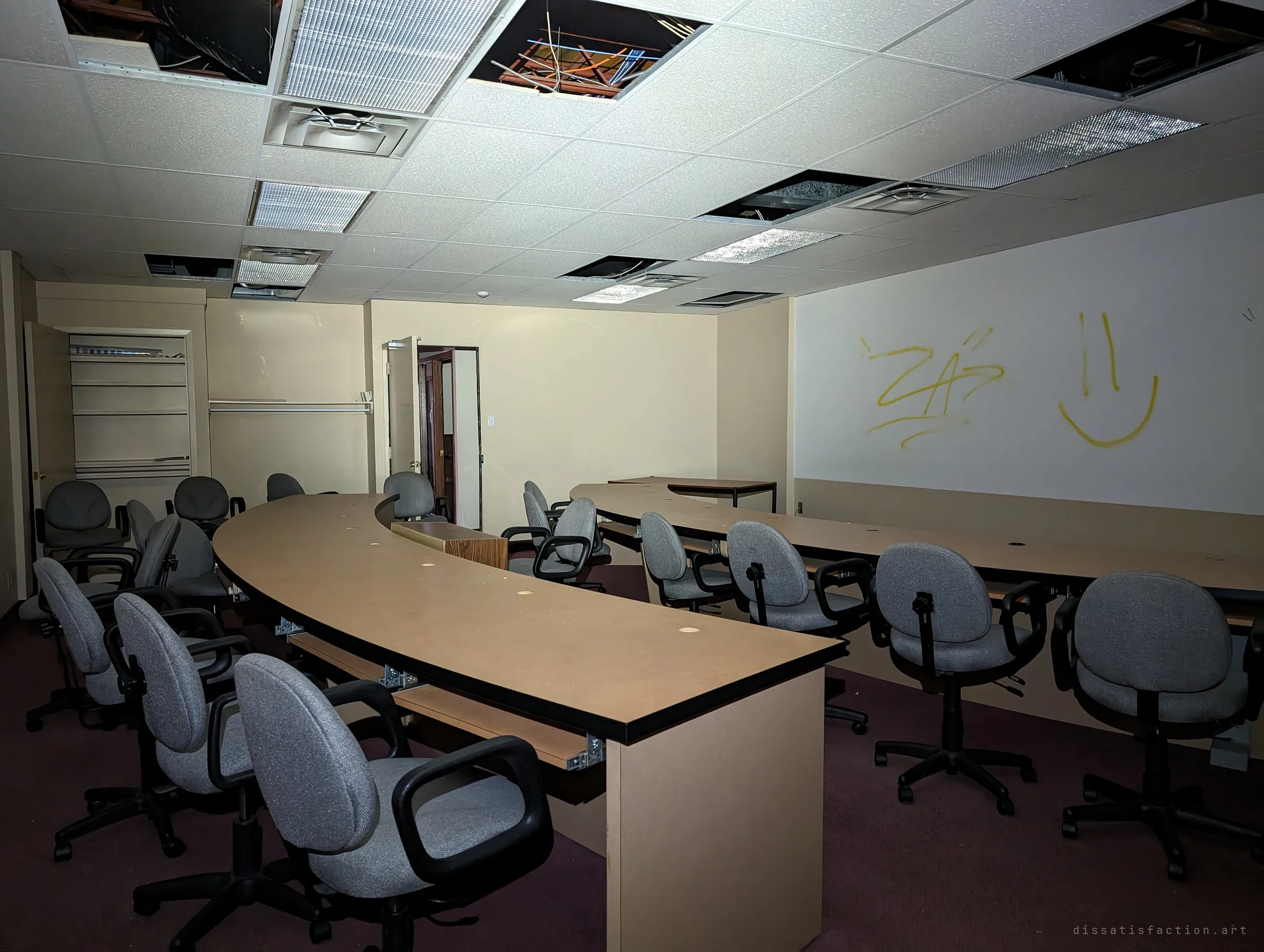 A training room with curved desks and rows of grey office chairs sits under a ceiling with missing tiles and dangling cables. Yellow spray-painted scribbles and a smiley face mark the wall.