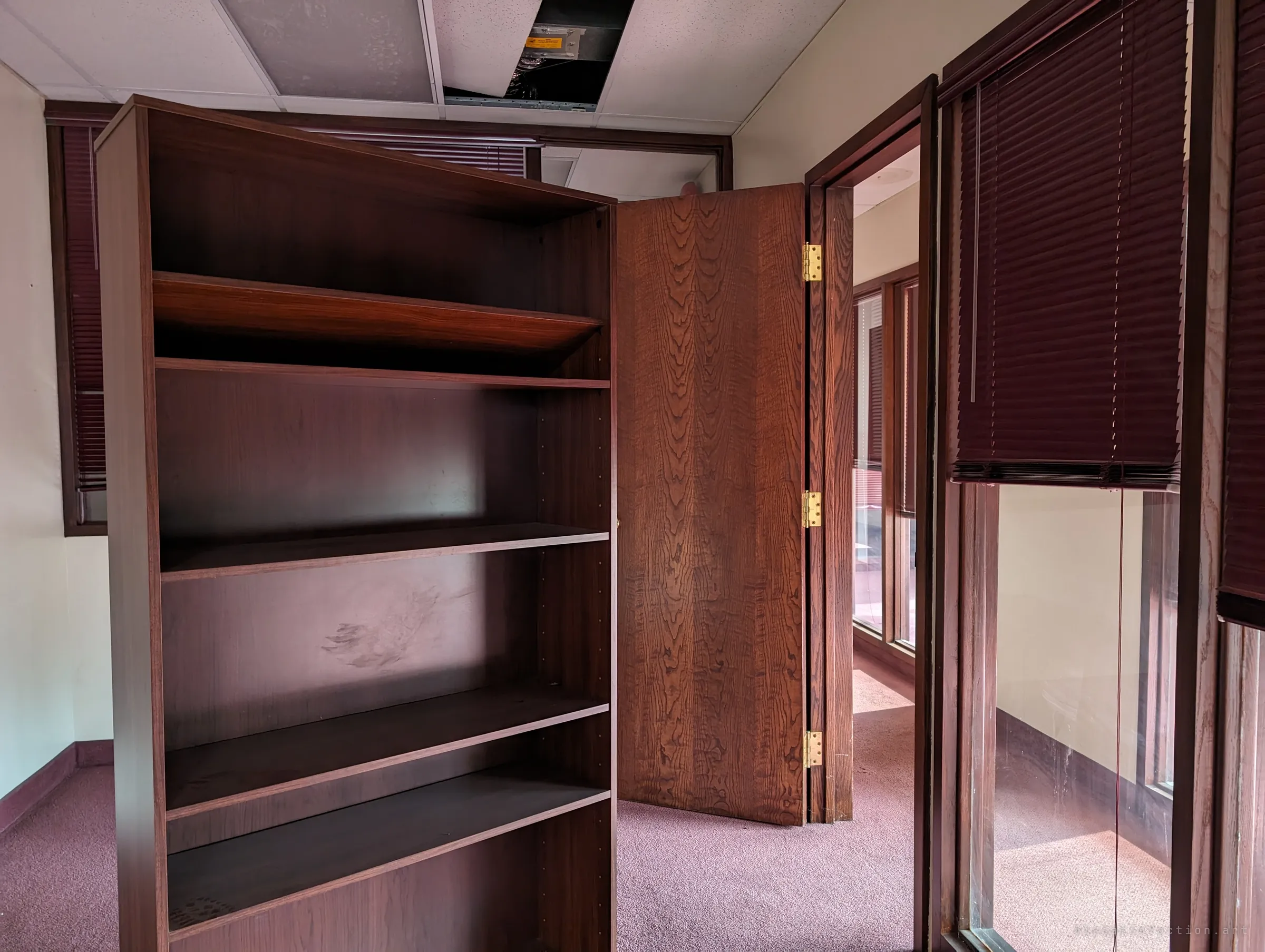 An empty bookcase stands in the middle of a small office beside an open folding door, maroon blinds half drawn and a ceiling tile missing above. The shelves are bare, the room already stripped of its paperwork.