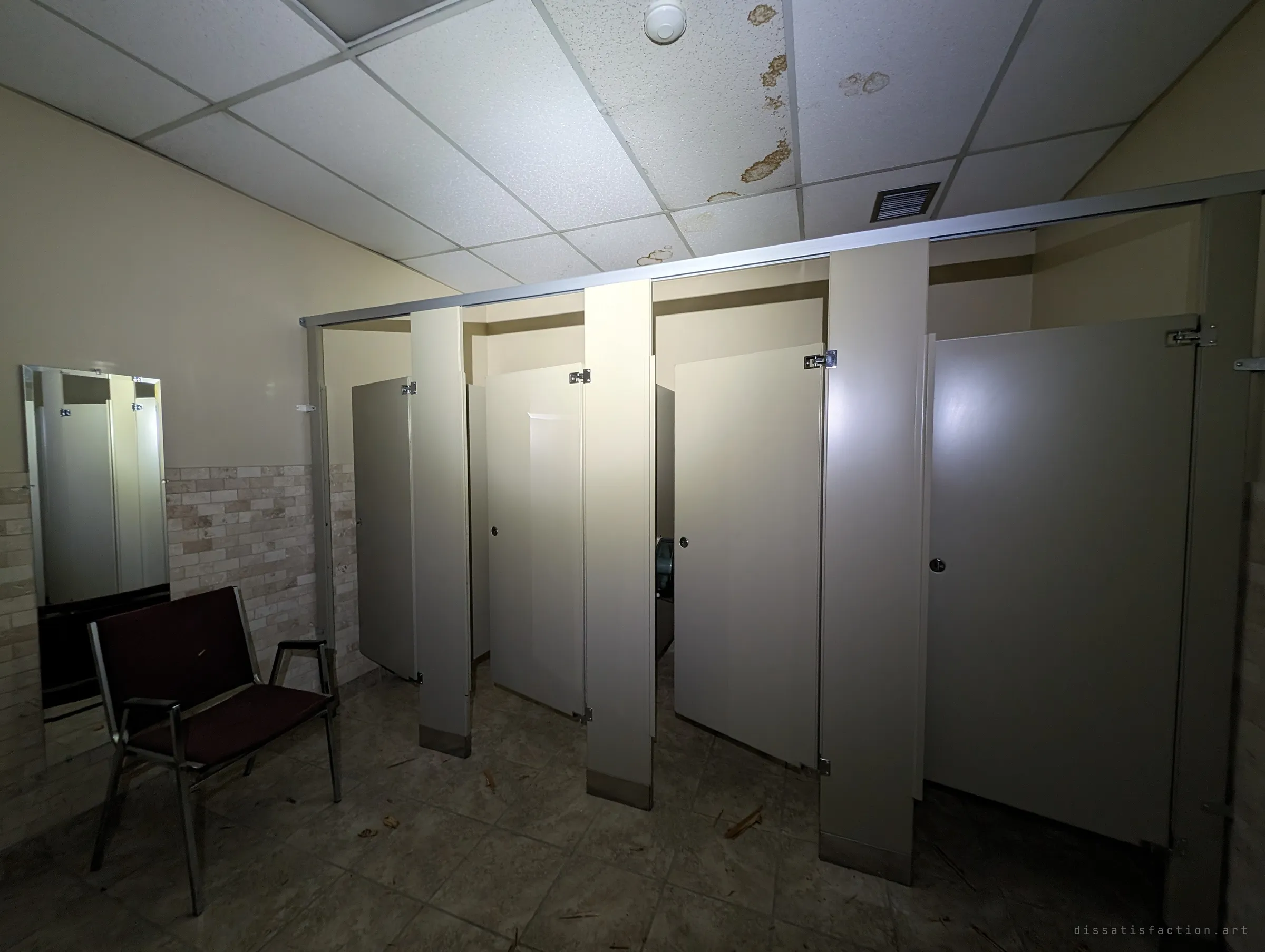 A row of beige toilet stalls with doors ajar under a water-stained drop ceiling, a single chair and a tall mirror by the tiled wall. Flashlight glare and leaves on the floor show how long the washroom has sat unused.