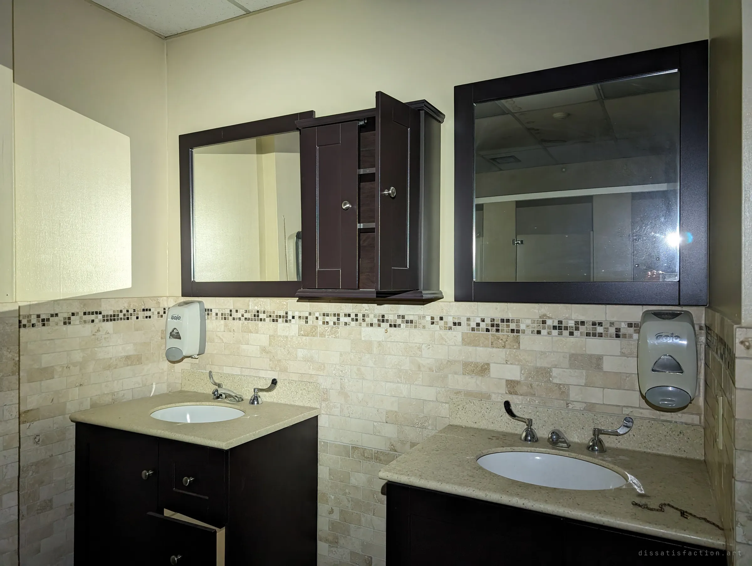 Two vanities with dark wood cabinets and framed mirrors over a travertine tile backsplash, soap dispensers still mounted and a medicine cabinet hanging open. The tidy finish contrasts with the dust and the empty room reflected behind.