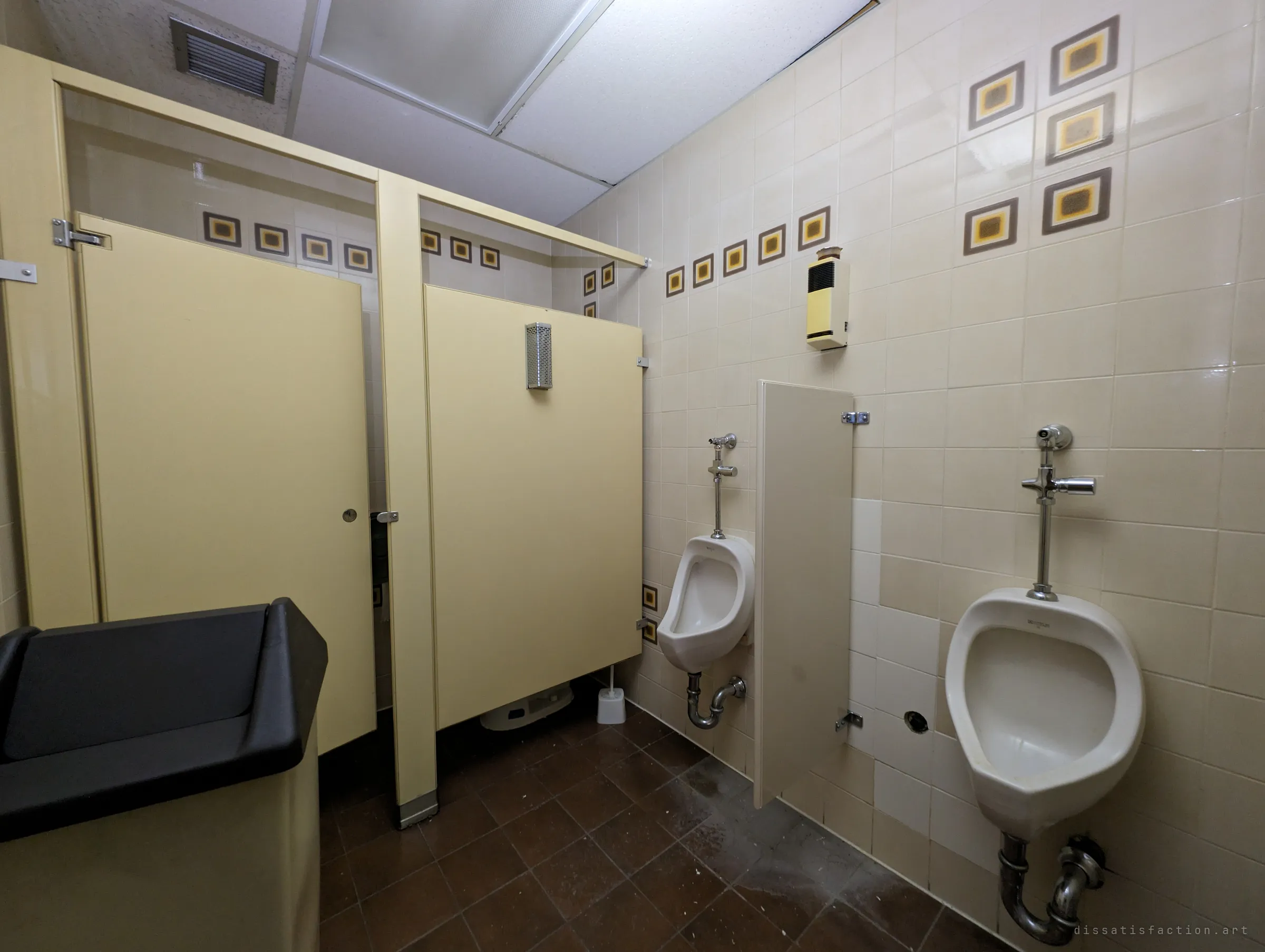 A men's washroom with butter-yellow stalls, two urinals and a wall of cream tiles set with retro brown-and-mustard square accents. The seventies décor has survived intact while the building emptied around it.