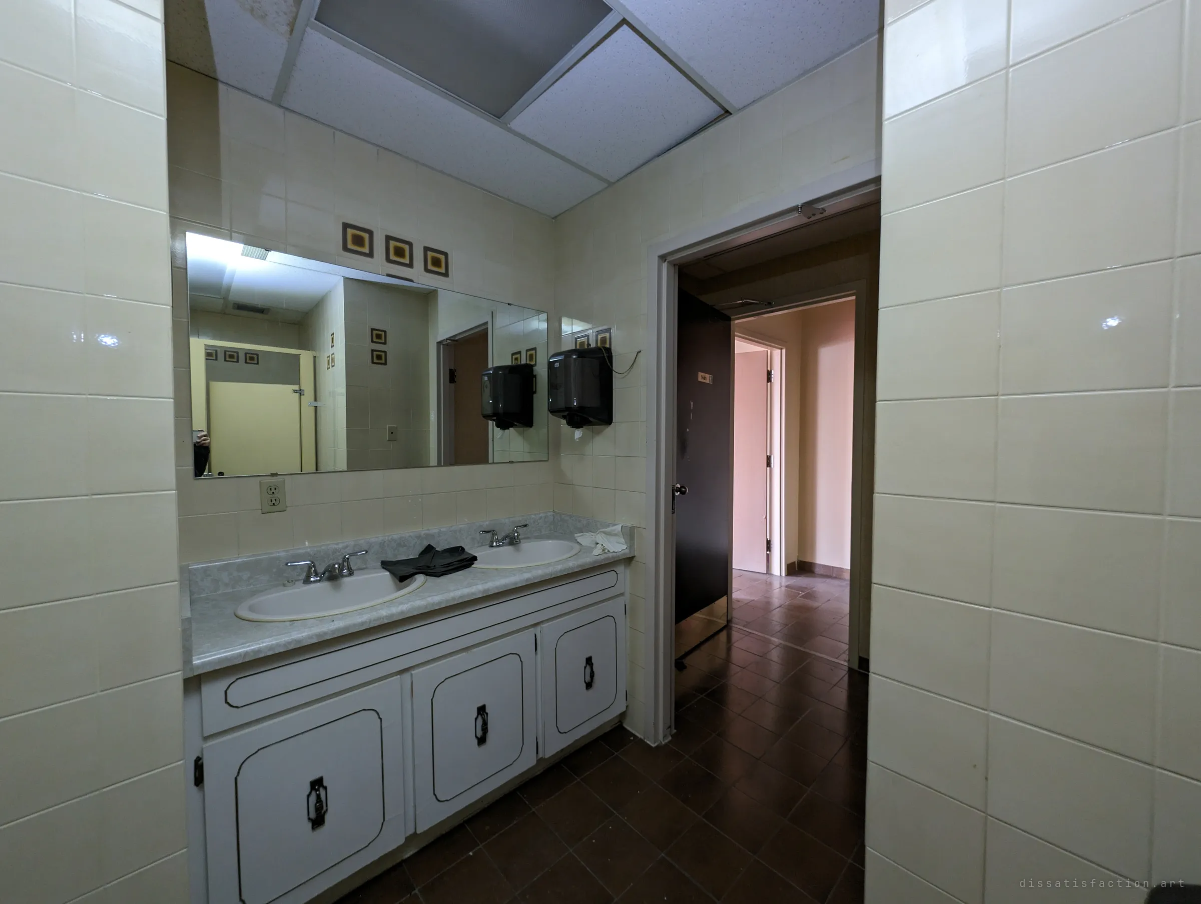 A washroom vanity with twin sinks, a long mirror and a pair of rubber gloves left on the counter, the doorway opening onto a dark-tiled corridor. The mirror catches the stalls and patterned tiles behind the photographer.