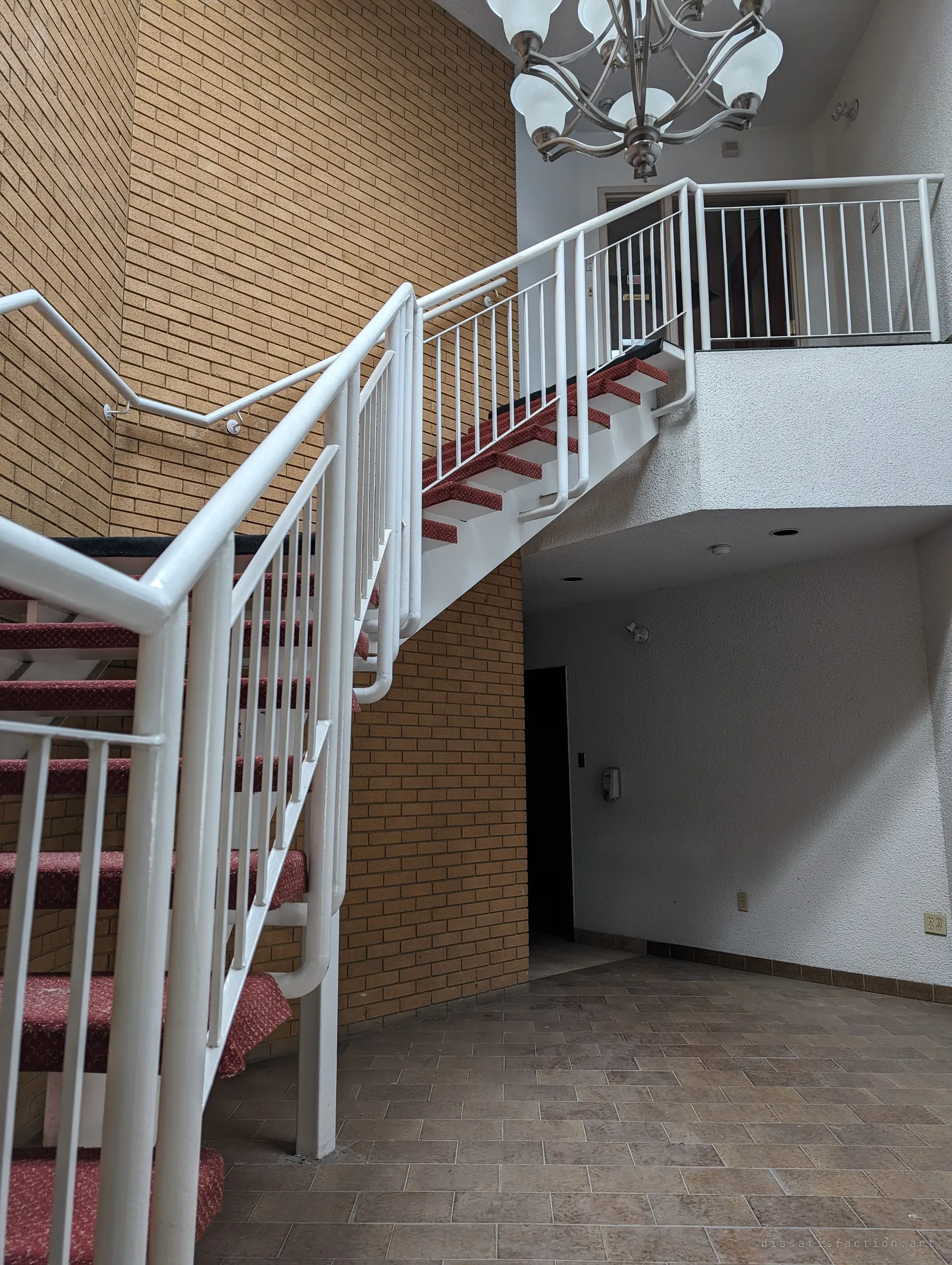 Looking up a white steel staircase with red carpeted treads in a tall brick atrium, a chandelier hanging over the landing. The lobby's once-grand entrance now leads to empty upper offices.