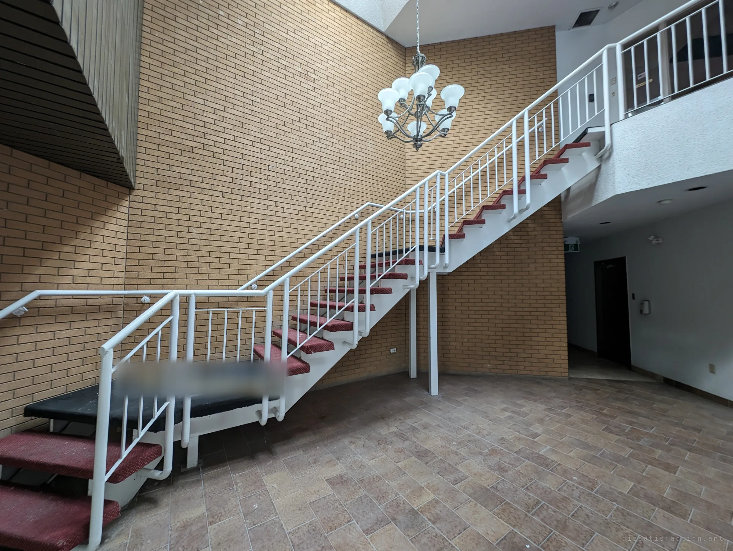 The full sweep of the atrium stair, white railings zigzagging up a two-storey buff brick wall past a hanging chandelier to a balcony above. The lobby floor below is bare and dusty.
