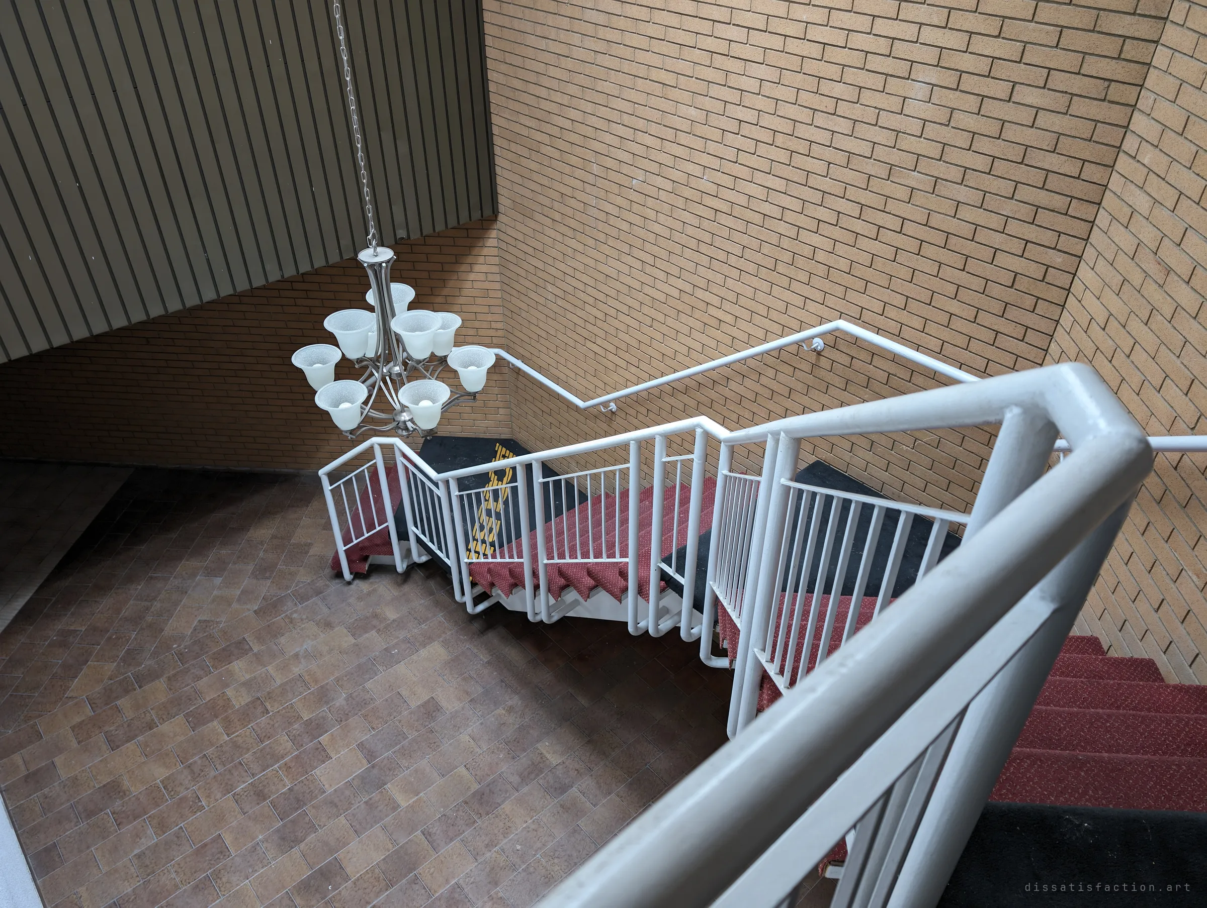 Looking down from the balcony over the white railing onto the red-carpeted stair and a chandelier suspended above the brick-tiled lobby. The height and the empty floor below give the atrium a hollow, echoing feel.