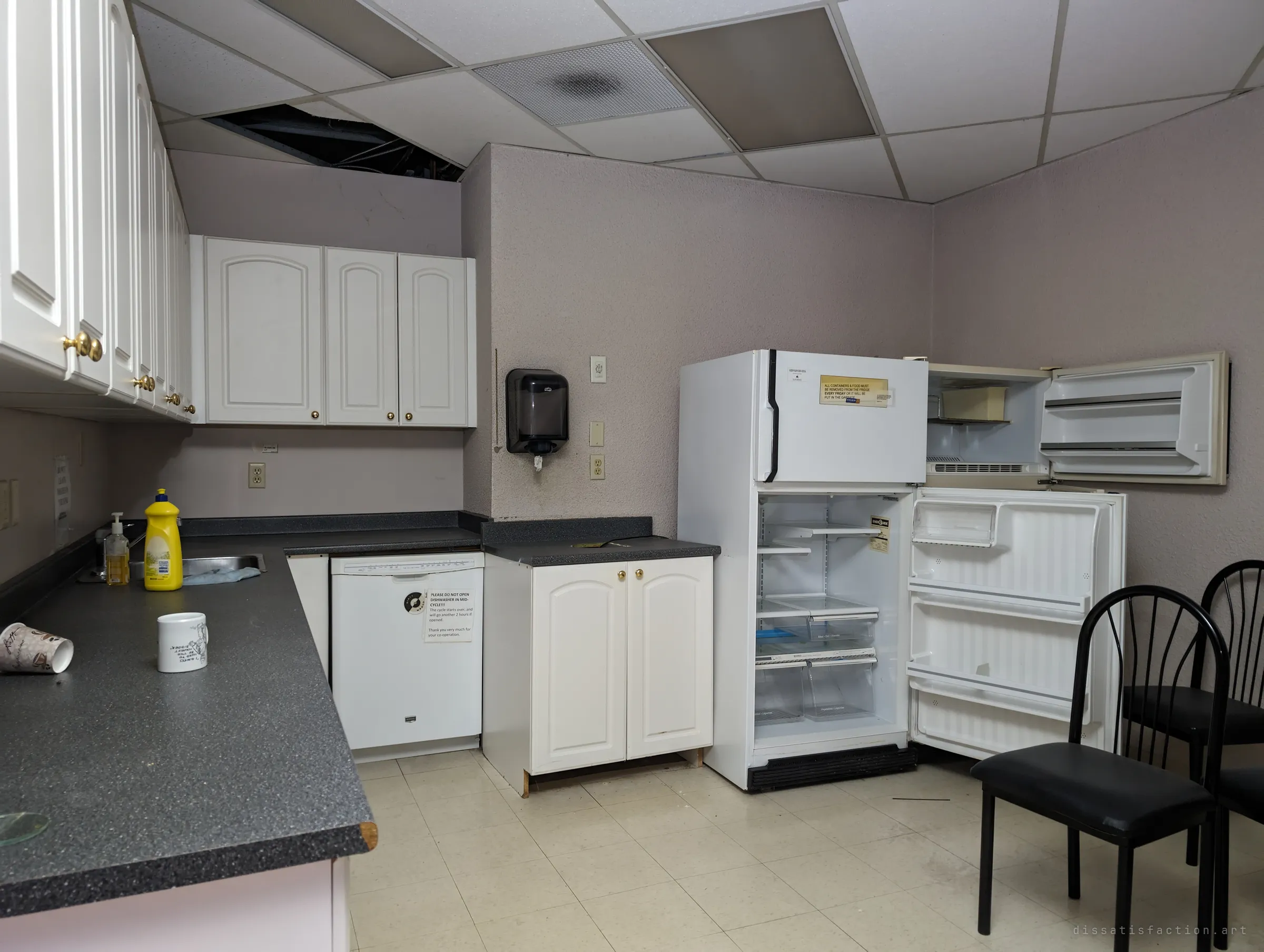 An office kitchenette with white cabinets, a dishwasher still carrying a polite do-not-open note and an old fridge standing open and empty. A mug and paper cup left on the counter hint at the last coffee break before the staff left.