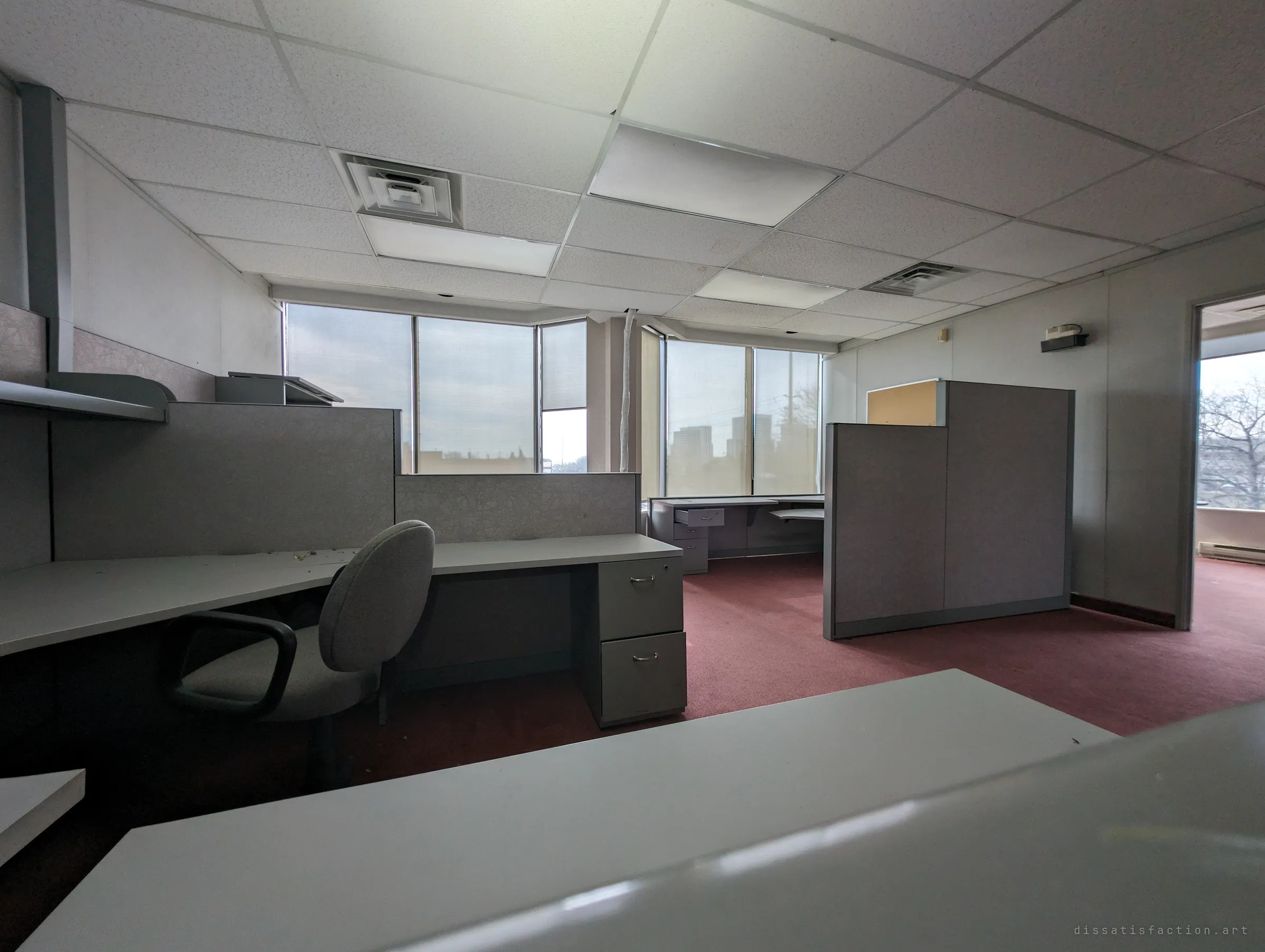 A wide view across an empty corner office, a lone chair pulled up to a bare desk, with pale daylight coming through tall windows. The furniture is still in place as if the staff just left.