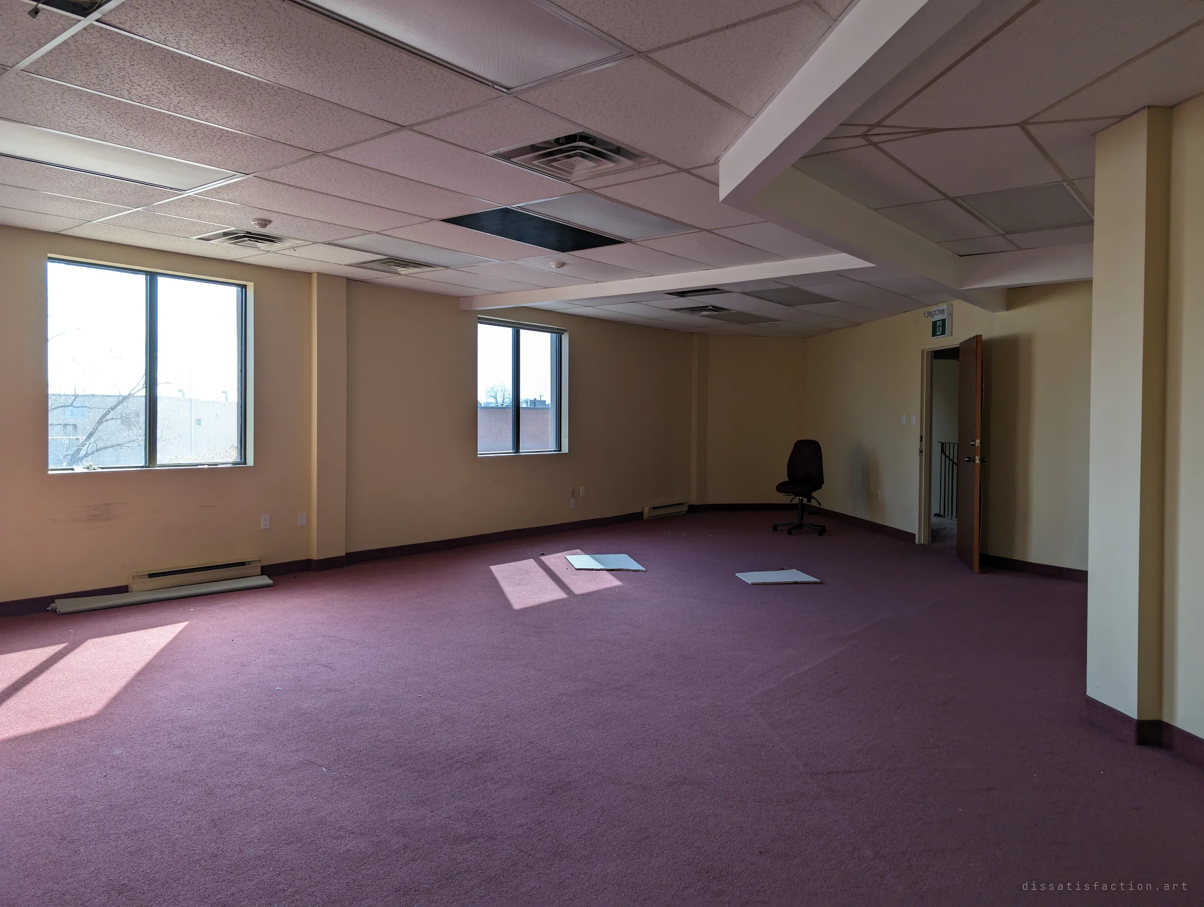 A large empty room with plum carpet, yellow walls and a single office chair left facing the corner. Sunlight falls through the windows onto a couple of fallen ceiling tiles.