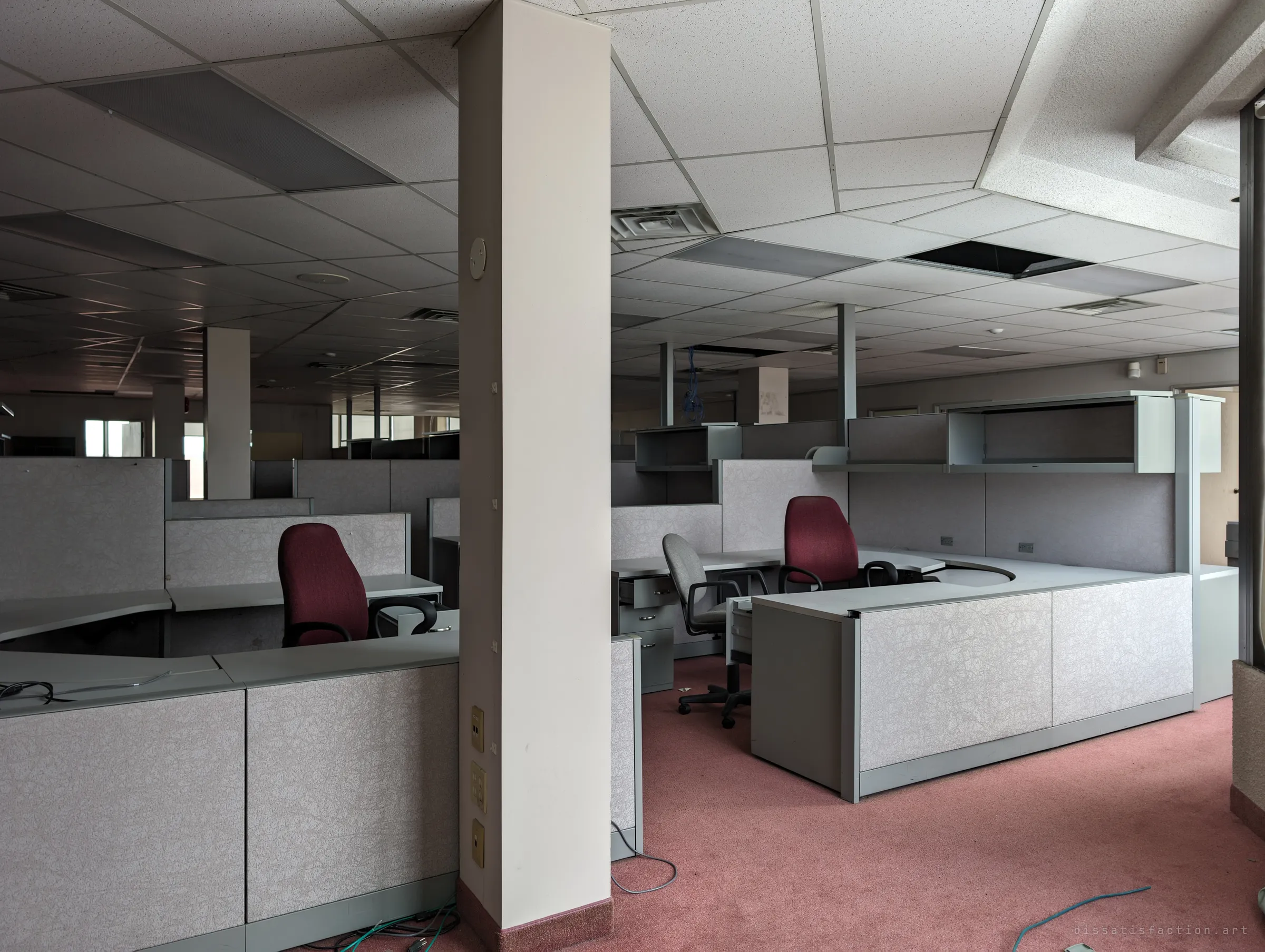 An open-plan office of grey cubicles with red chairs, split by a white column, with a few ceiling tiles missing. The floor runs back into darkness, rows of workstations left in place.