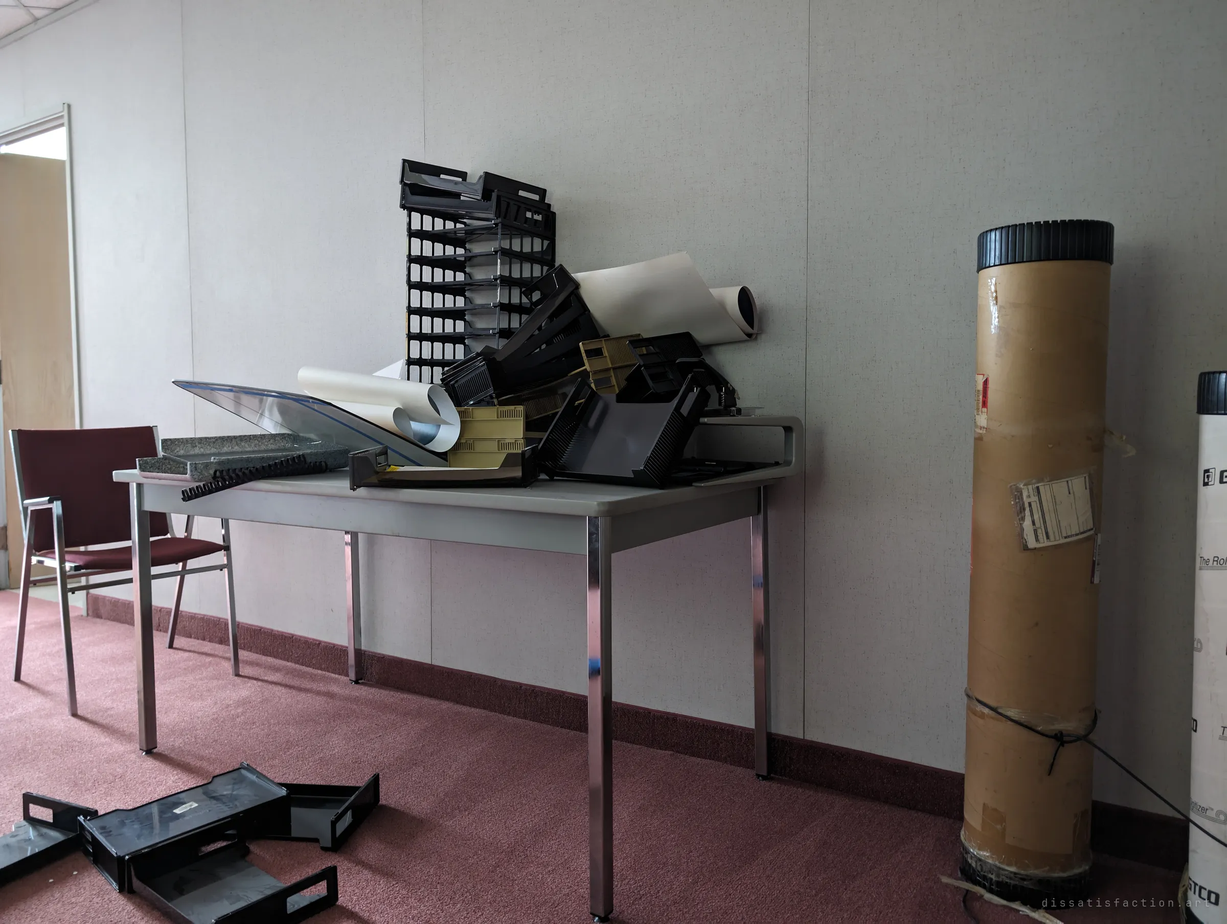A folding table piled with black desk trays, rolled plotter paper and binding combs, next to tall shipping tubes leaning against the wall. The clutter looks like the last things someone meant to take away.