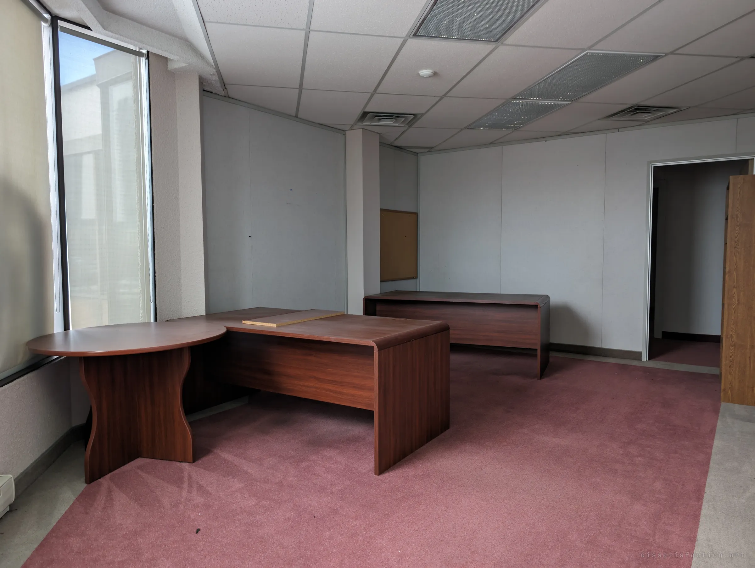 An empty executive office with matching wood-grain desks and a round meeting table on faded pink carpet. A cork board hangs bare on the wall.