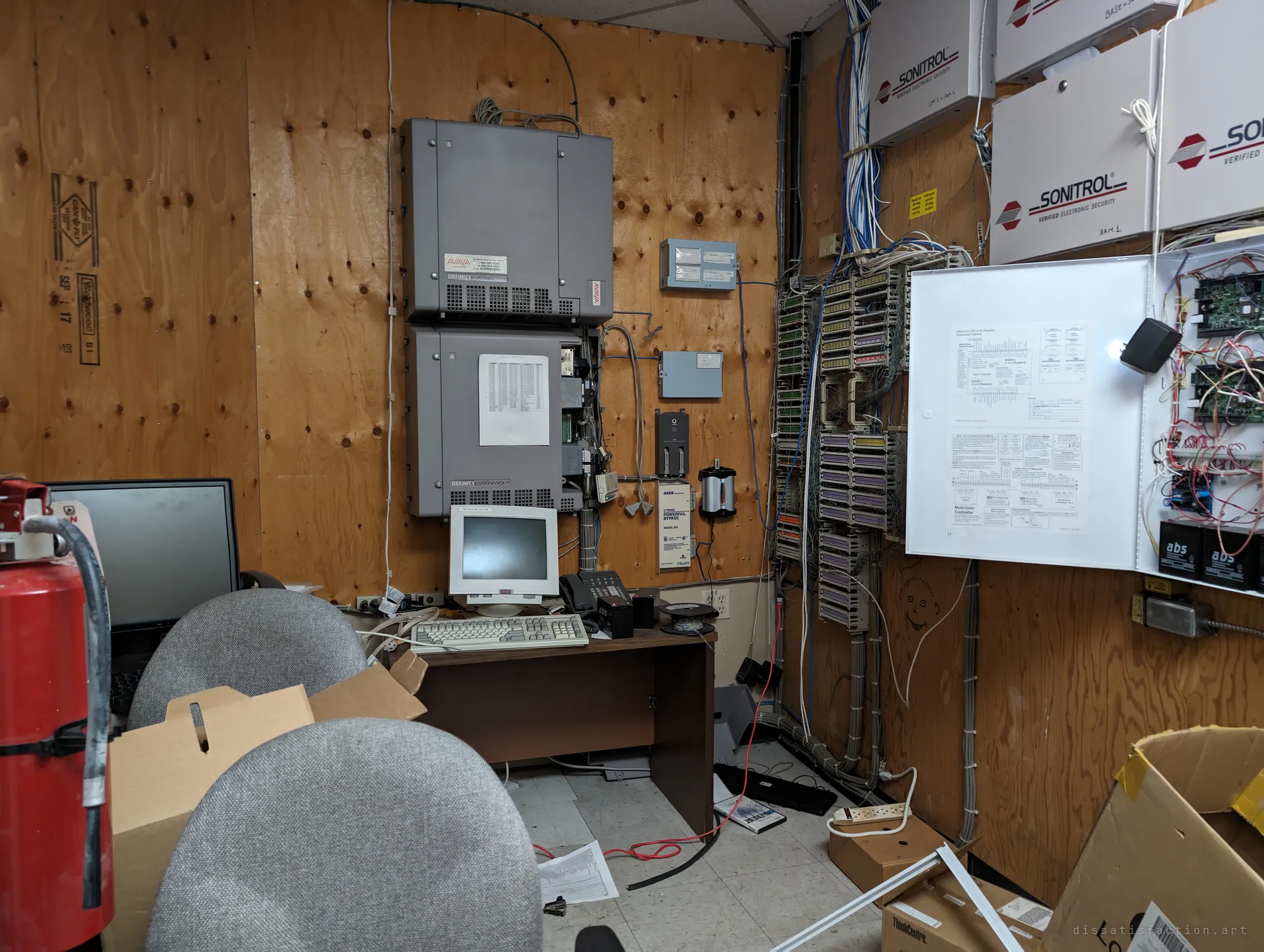 The building phone and security room, plywood walls hung with an old phone switch, punch-down blocks, alarm panels and tangled cabling, with a beige terminal on a desk. A small face has been doodled on the wall among the wires.