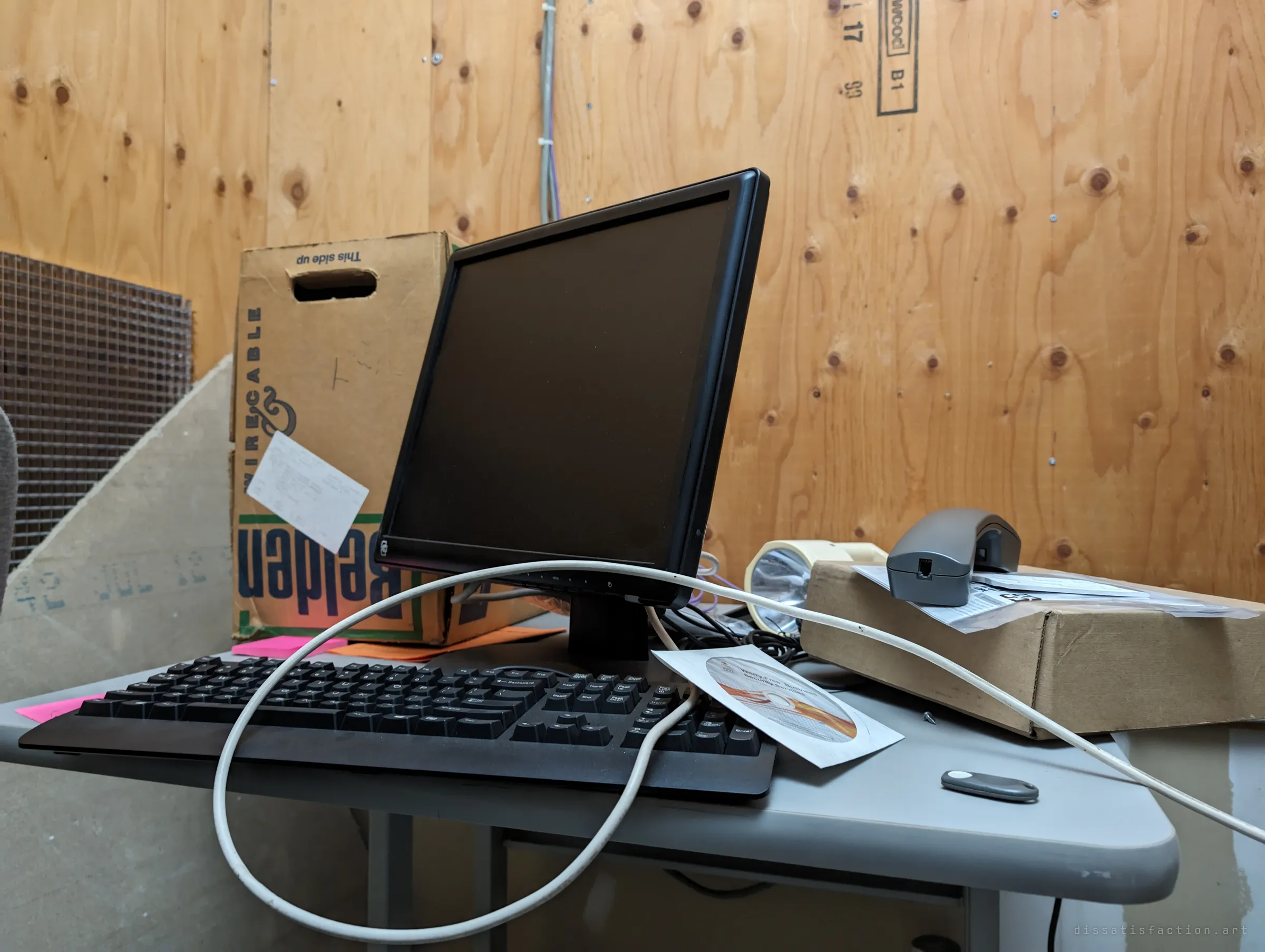 A flat-screen monitor and keyboard left on a grey desk, a cable looped over them and a software disc beside them, in front of a plywood wall. Sticky notes and a cable box sit where the last technician left them.