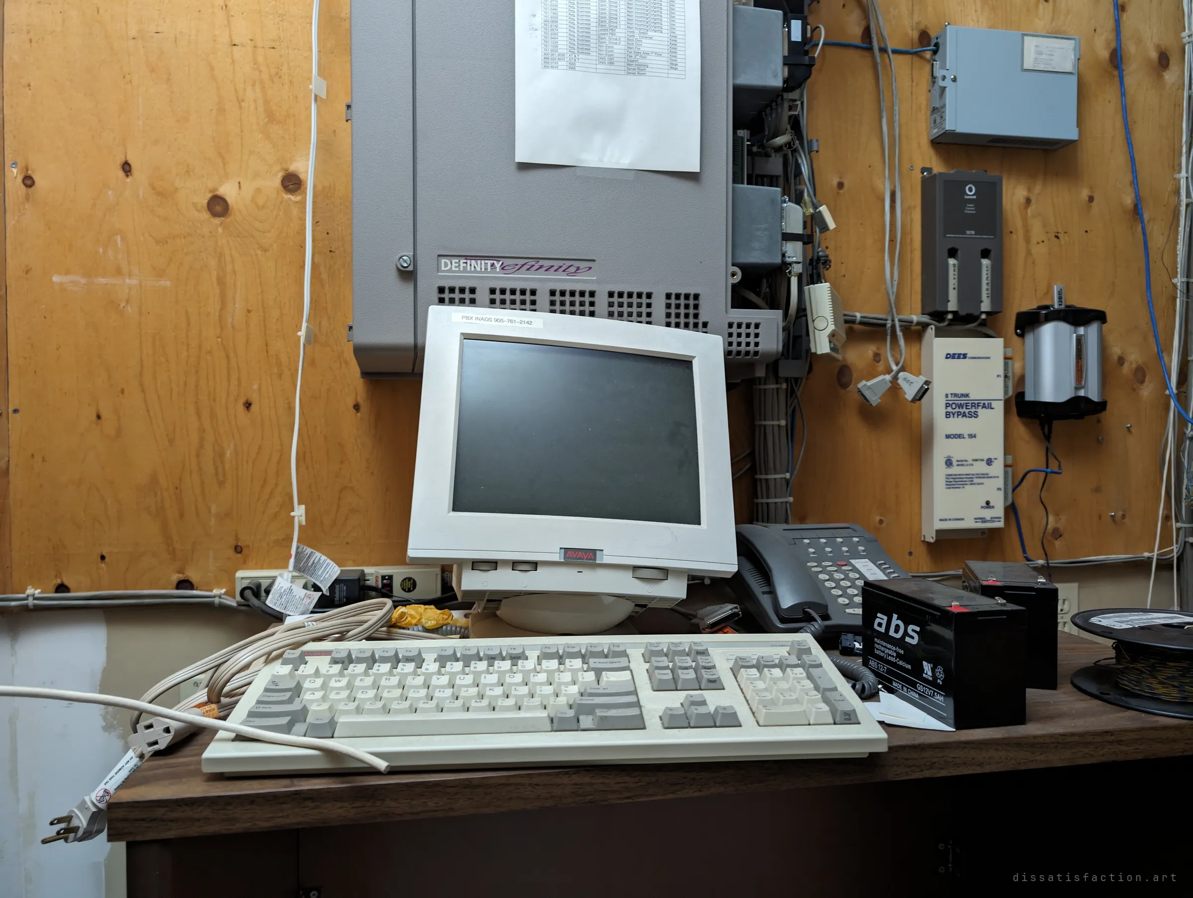 A close look at the old phone-switch terminal: a yellowed CRT monitor and a beige keyboard under the grey cabinet, with a backup battery and a desk phone beside them. Technology from another era, left running until the lights went out.