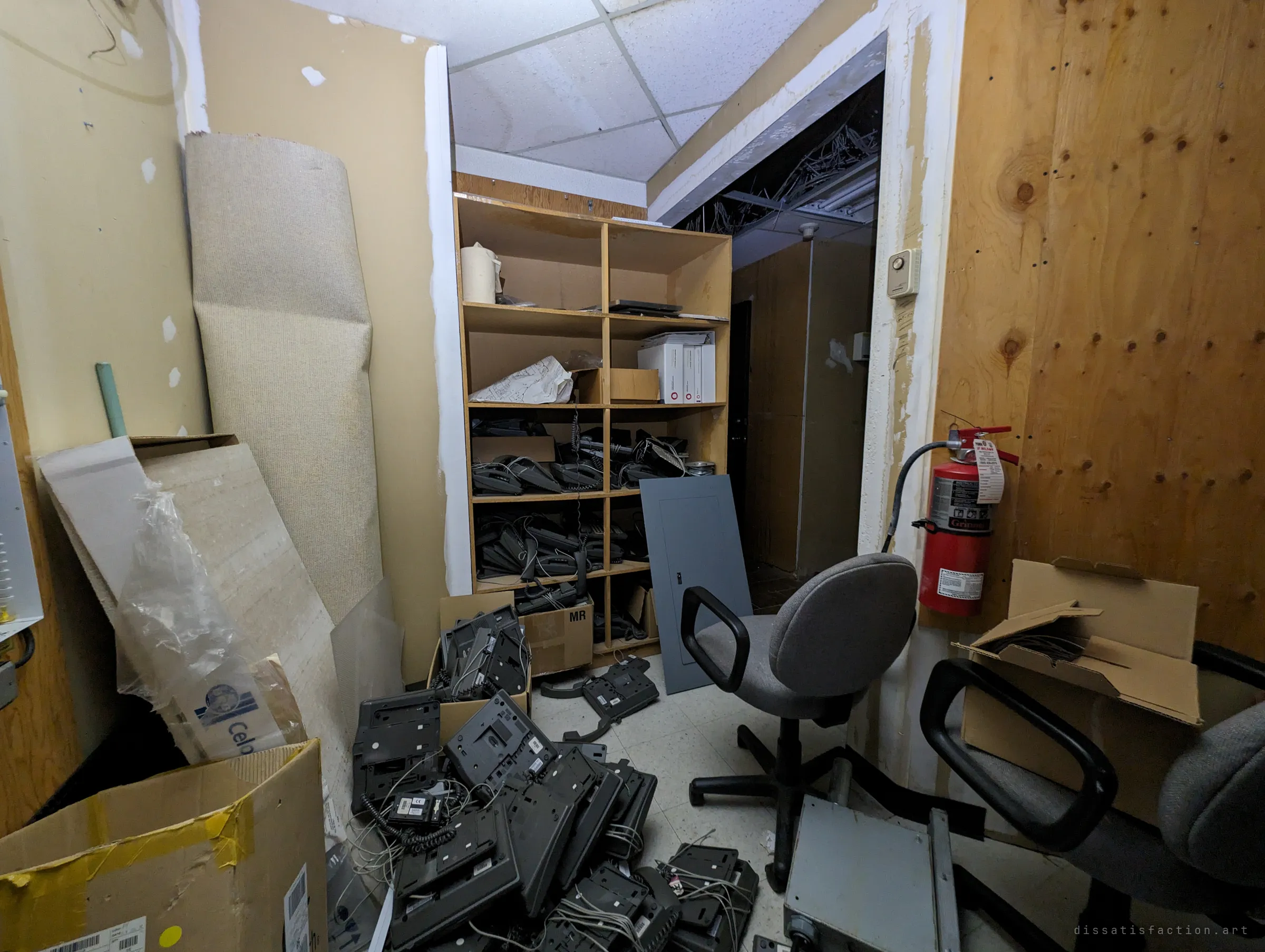 A cramped back room piled with discarded desk phones, rolled carpet and office chairs, with a shelf of more handsets and an open ceiling spilling cables beyond. The office's phone system was ripped out and dumped here when the place emptied.