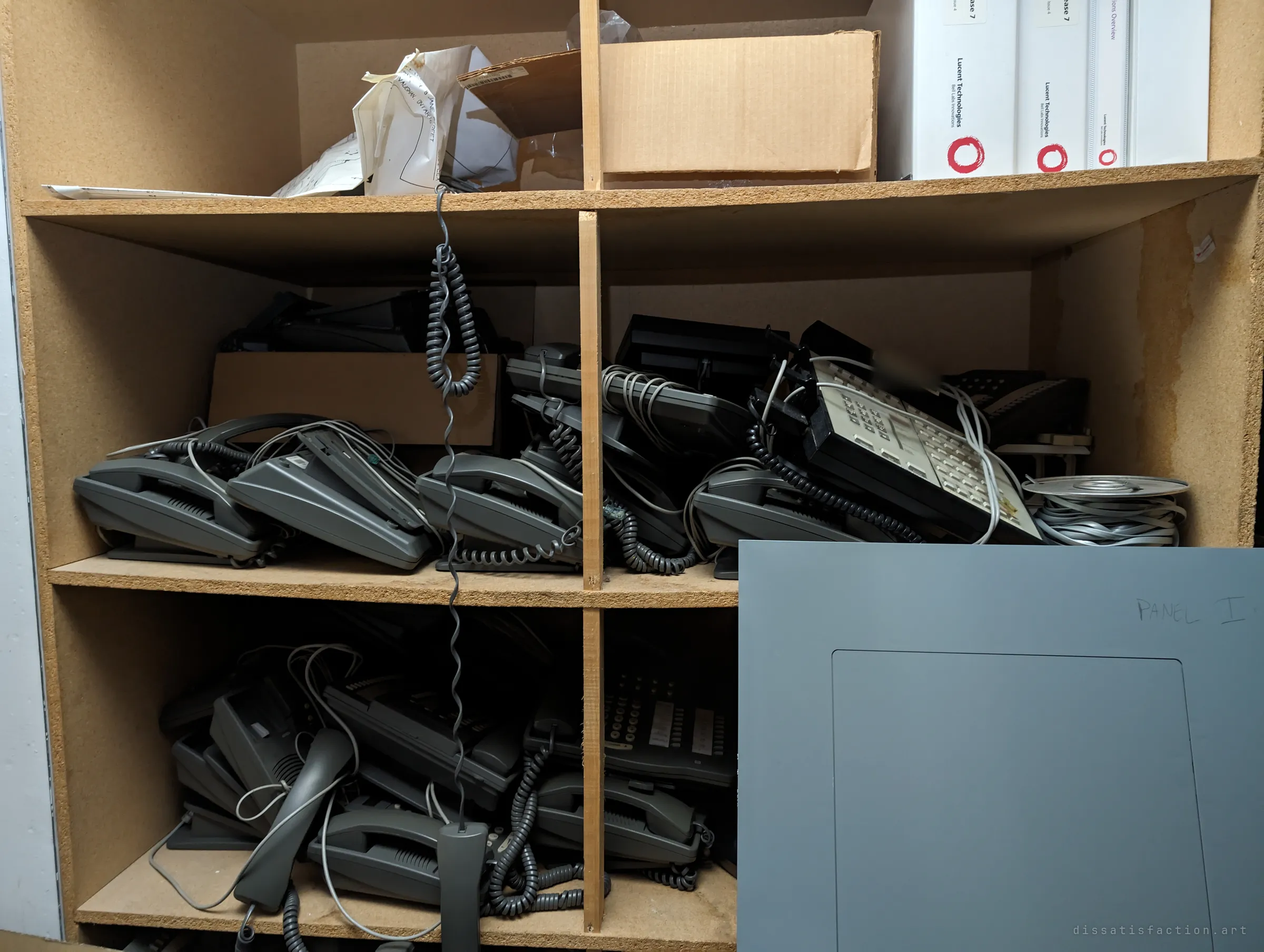 Shelves crammed with tangled grey desk phones and old telecom binders, a loose electrical panel cover leaning against them. Decades of office hardware left to gather dust.