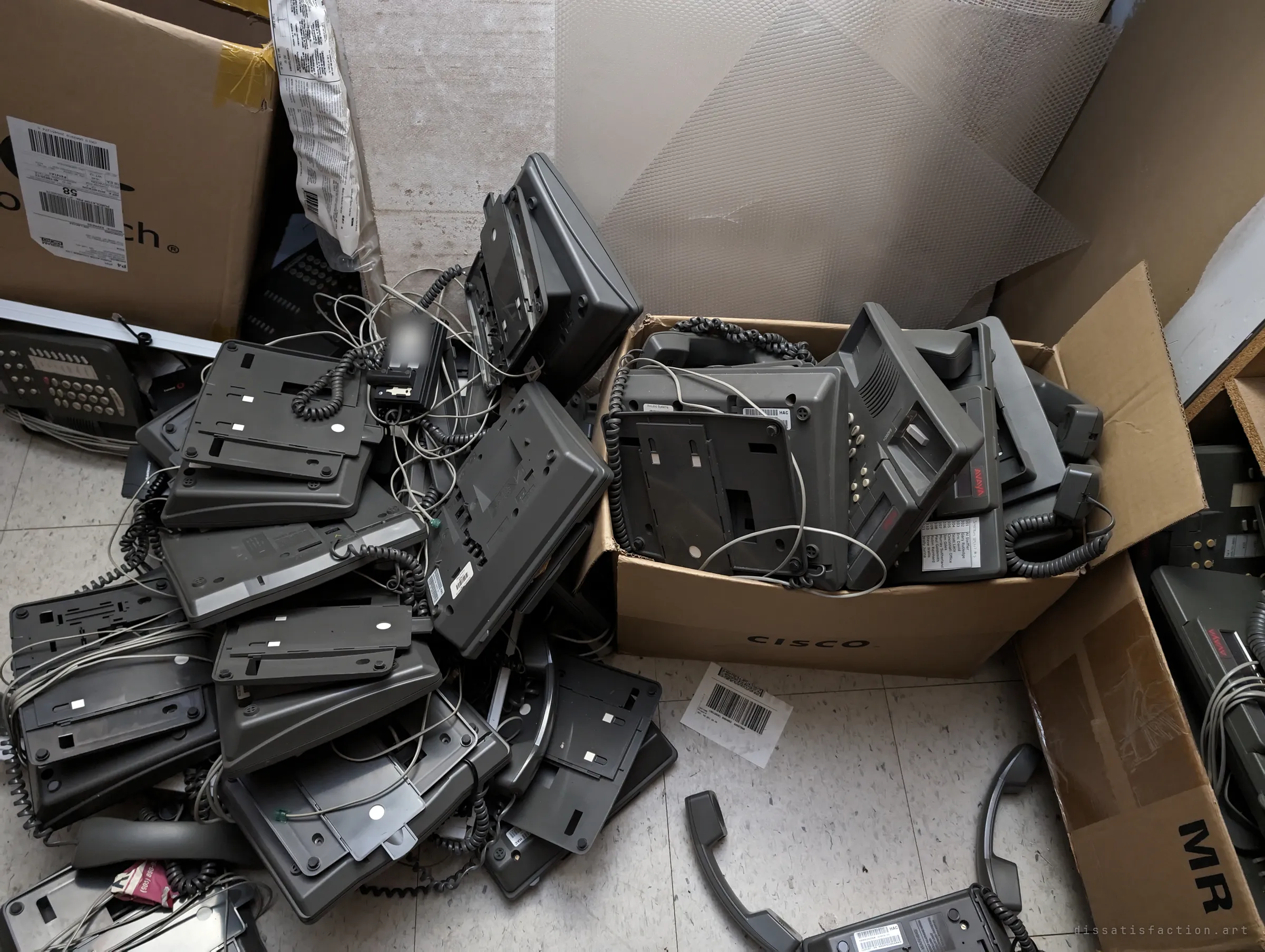 A heap of upturned desk phones and coiled cords spills across the tiled floor beside a cardboard box stuffed with more handsets. The whole phone system abandoned in a pile.