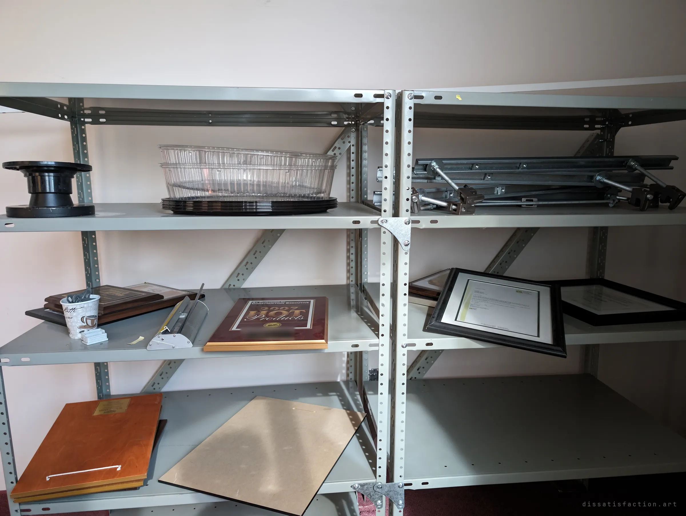 Grey steel shelving holding leftover awards, framed letters and spare hardware in a sunlit storeroom. The company's old honours left behind on the shelf.