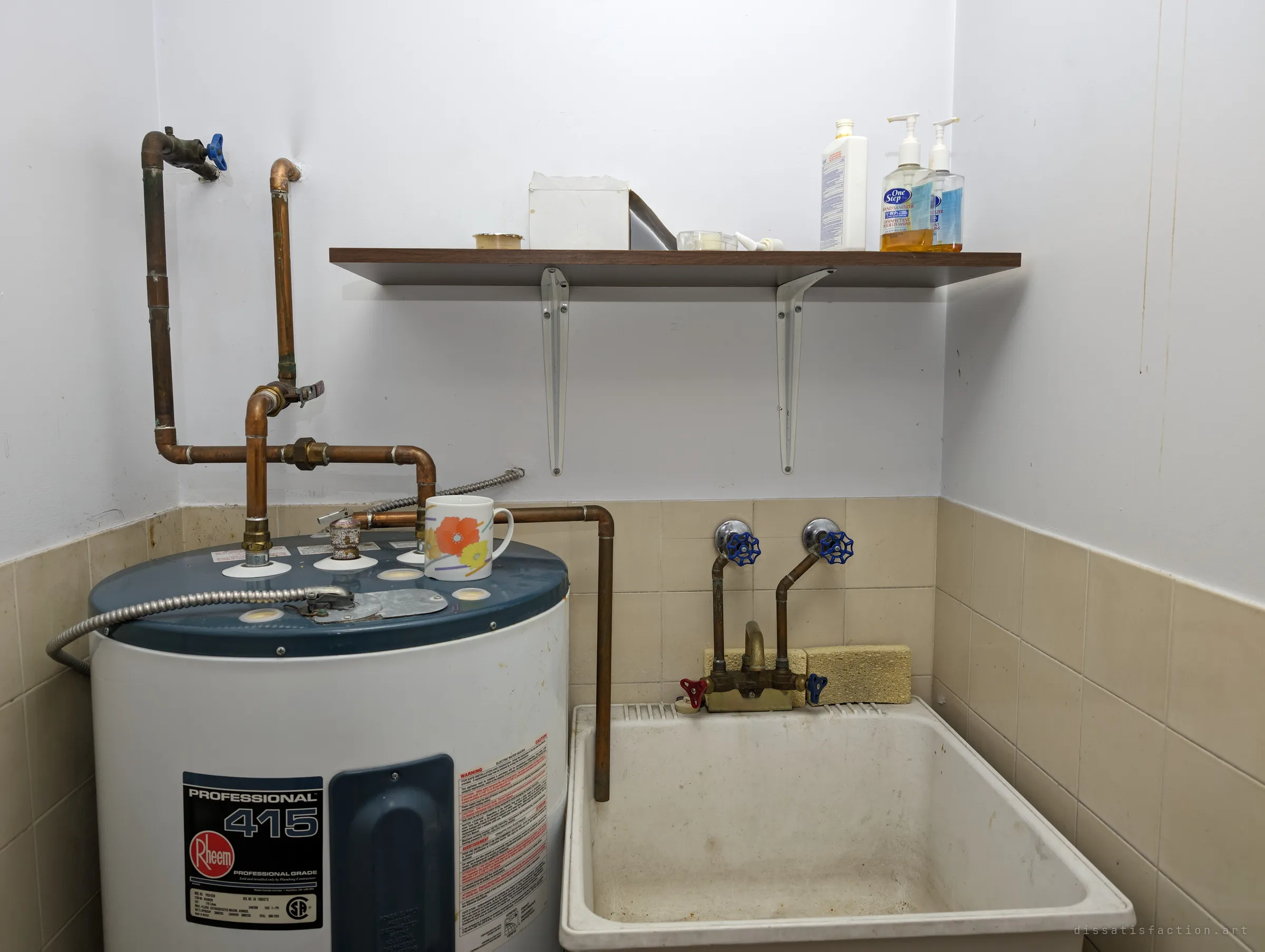 A small utility closet with an electric water heater, a utility sink and a shelf of hand sanitiser, a patterned mug still sitting on the tank. An everyday corner frozen as the staff left it.