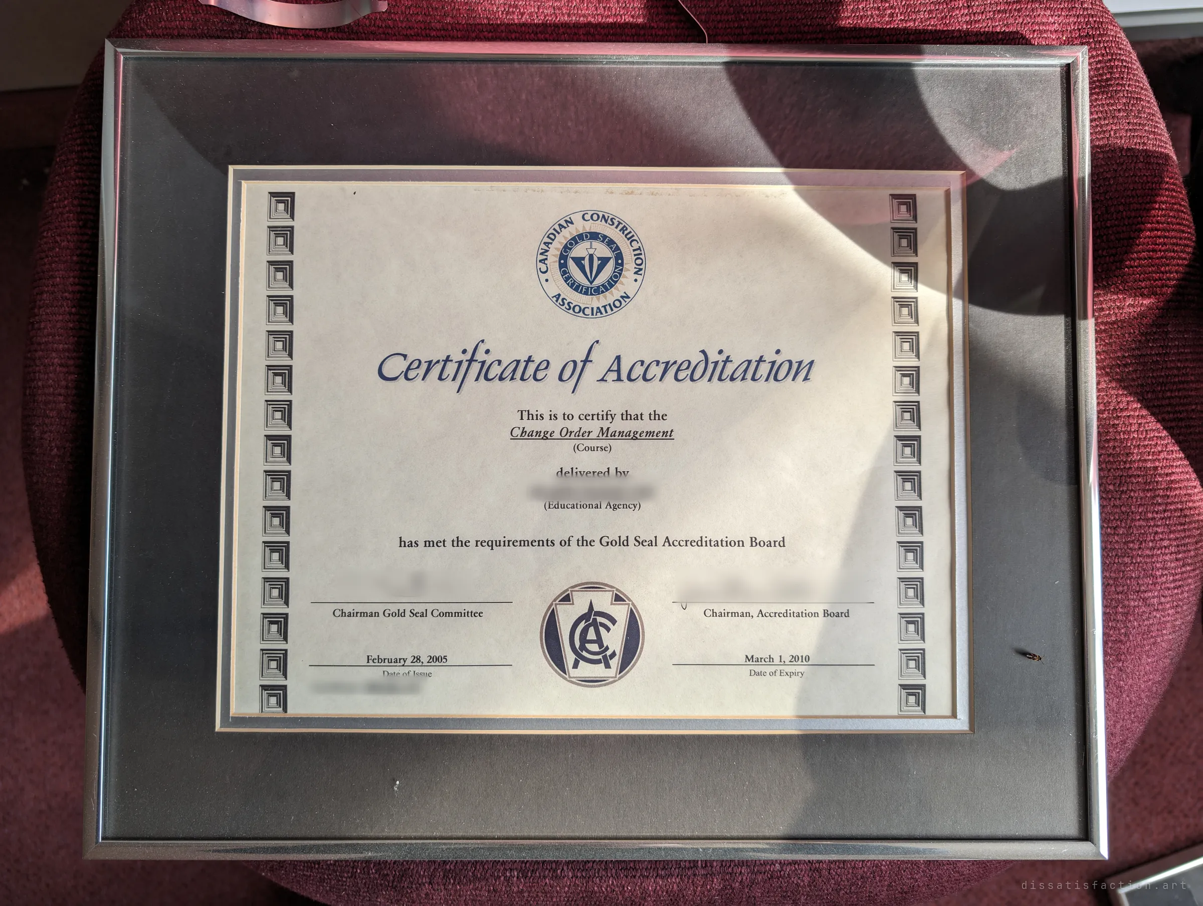 A framed accreditation certificate from a construction industry association, lying in sunlight on a worn red chair. Proof of the business's past standing, abandoned with everything else.