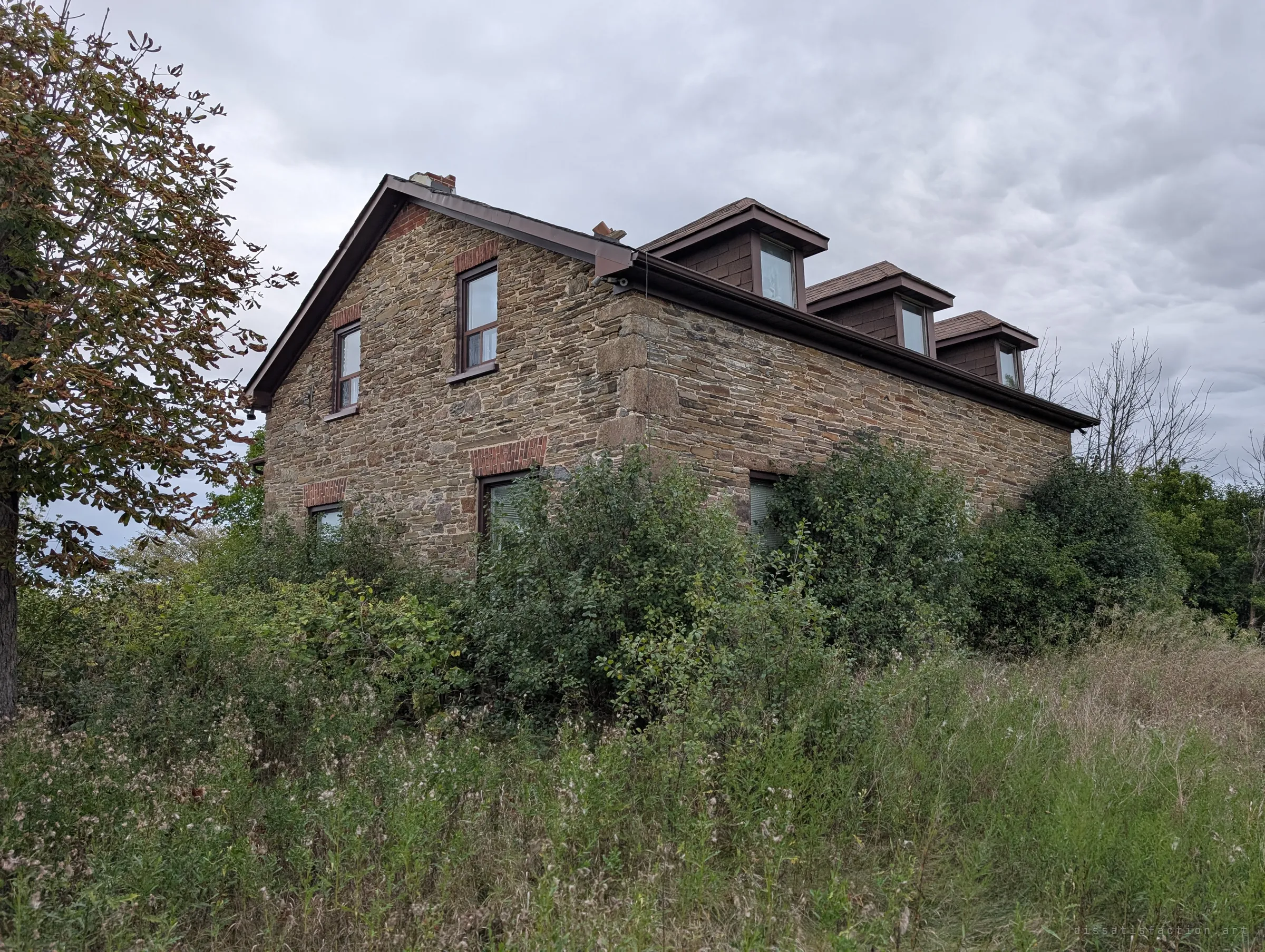 An overgrown fieldstone farmhouse with three dormer windows under a heavy grey sky, wild brush swallowing its ground floor. The opening shot of a house that nature is quietly taking back.