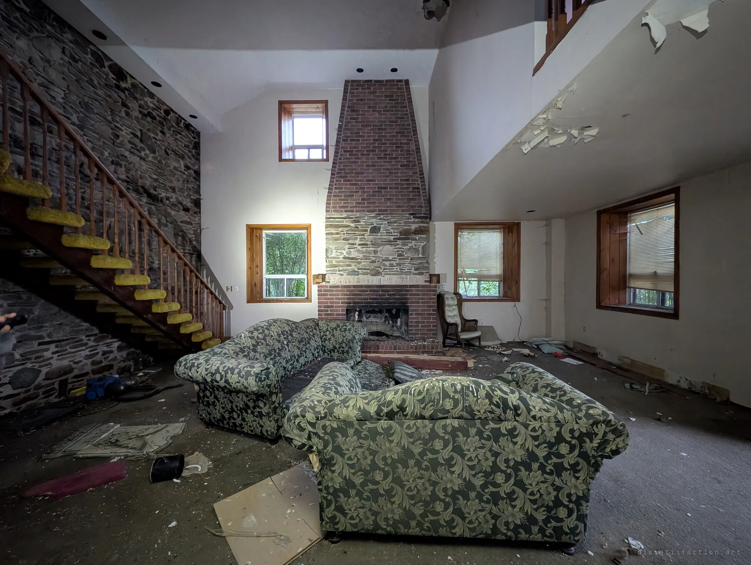 A double-height living room with a tall brick chimney, a stone wall with an open staircase and two floral sofas amid fallen plaster. The house still looks furnished, as if its owners simply walked away.