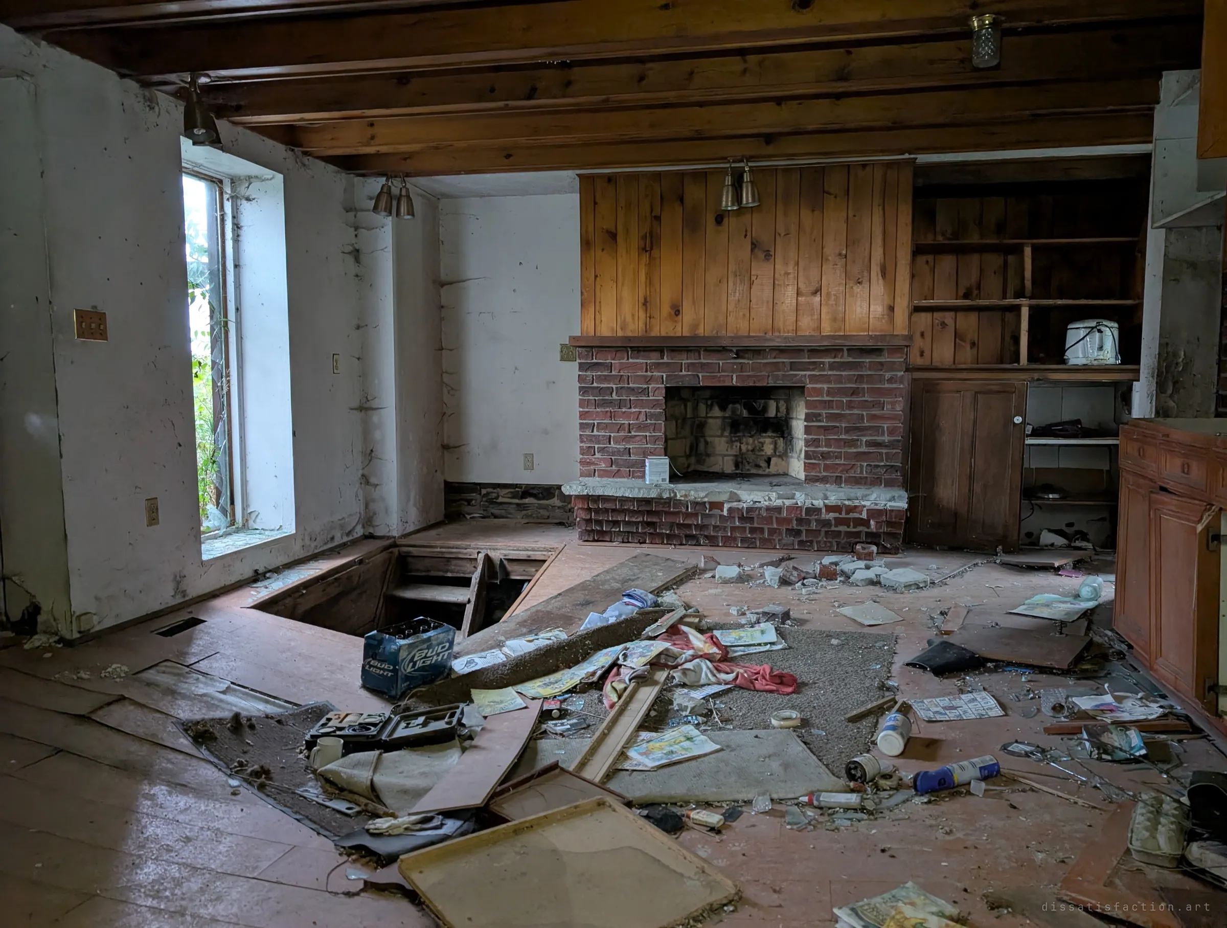 A beamed kitchen with a brick fireplace and pine panelling, the floor opened to a cellar hatch and strewn with comics, a beer case and debris. Everyday leftovers show how recently the house was lived in.