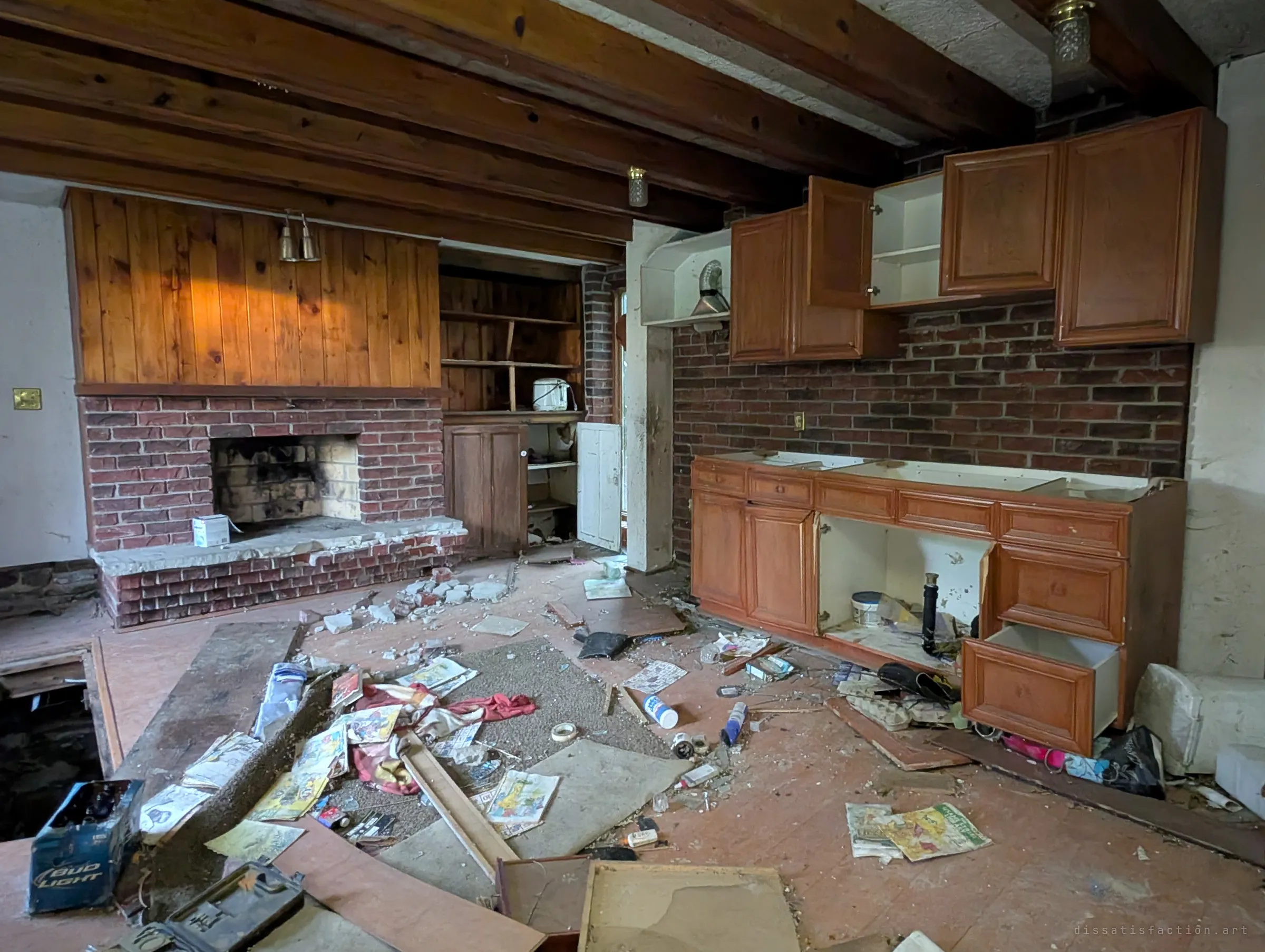 The same beamed kitchen from another angle, with open wooden cabinets against a brick wall and litter spread across the floor. The gutted sink cabinet and scattered belongings show the house being picked apart.